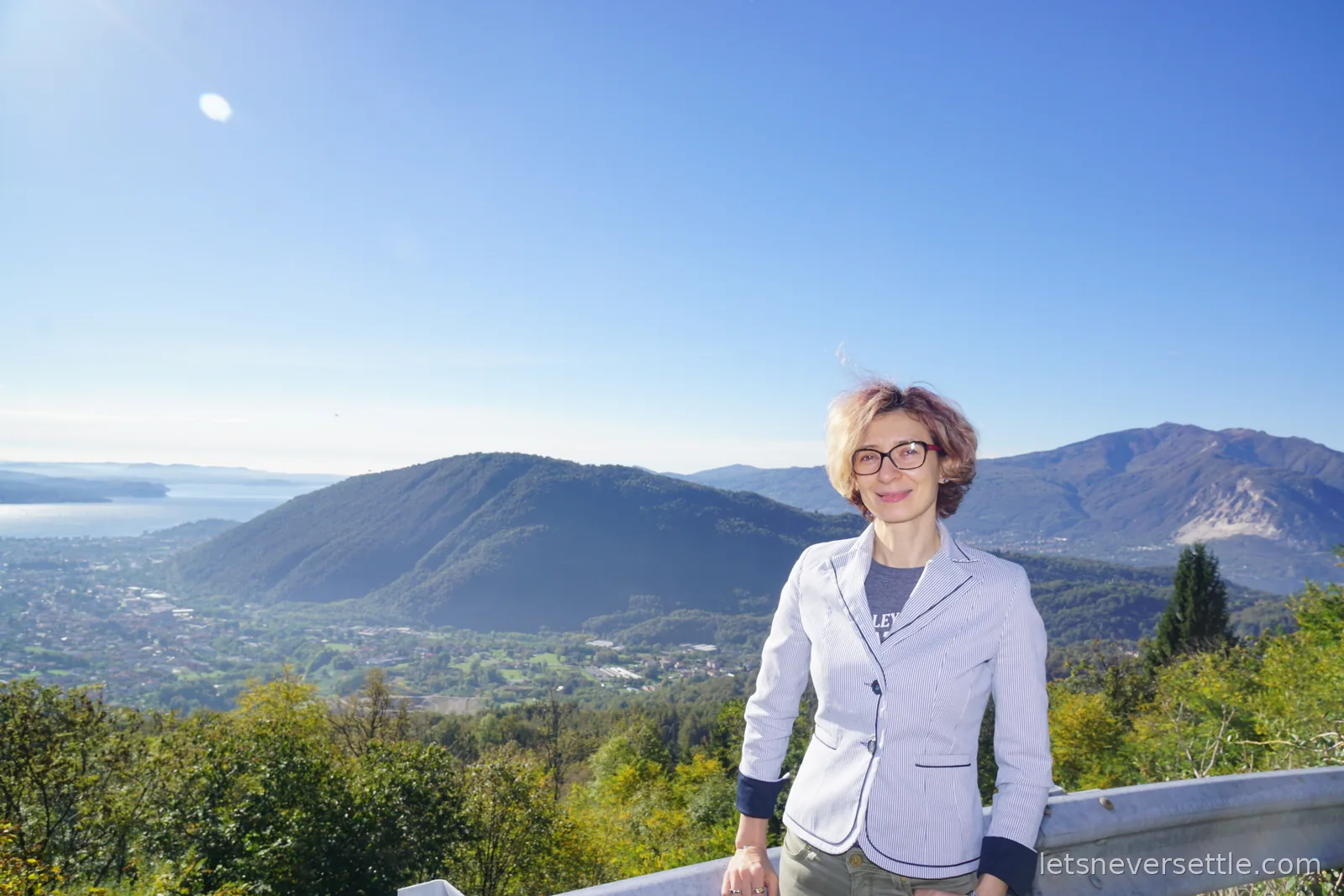 Inga in a striped blazer leaning on a crash barrier on a mountain road, with wooded ridges and Lake Maggiore spread out far below under a clear sky