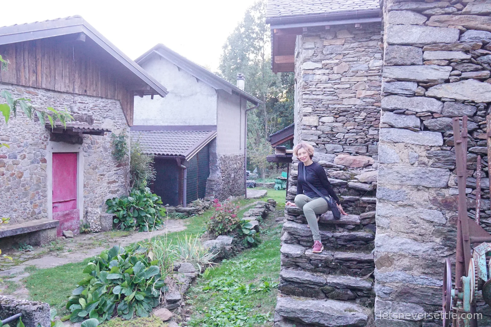 Inga sitting on rough stone steps built into a dry stone wall between old stone and timber barns, with a bright pink door on the barn opposite and a vegetable patch in front