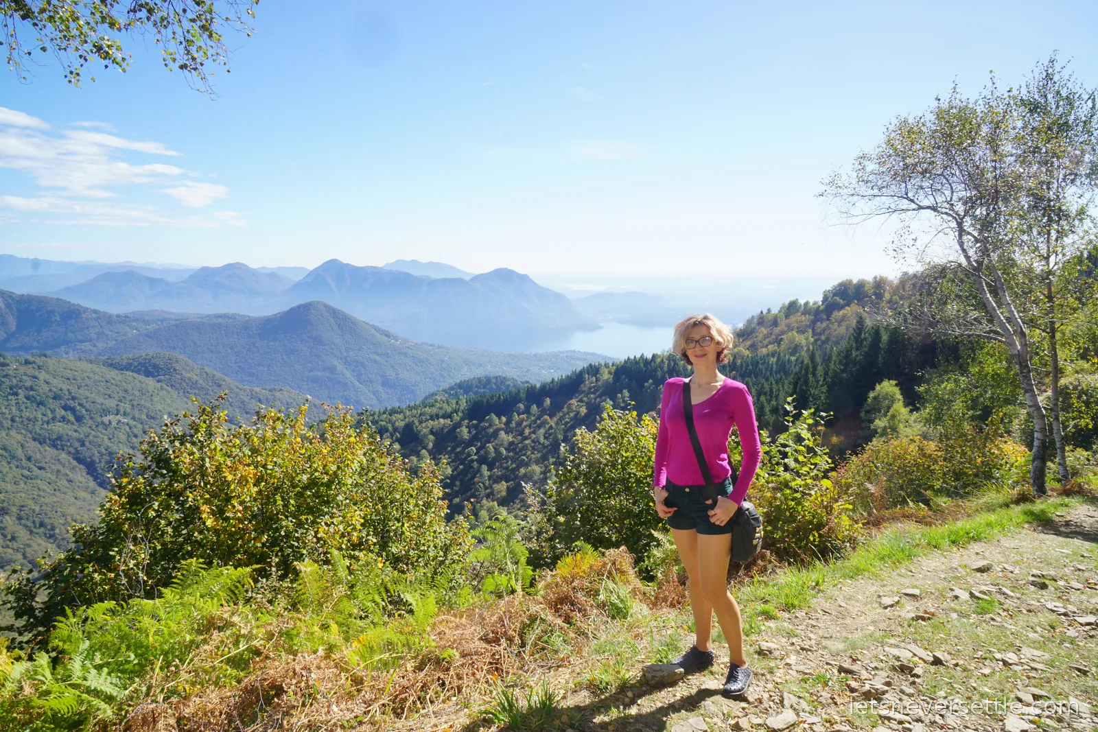 Inga in a bright pink top standing on a stony hillside path with a small bag, wooded ridges dropping away and a hazy arm of Lake Maggiore in the distance below
