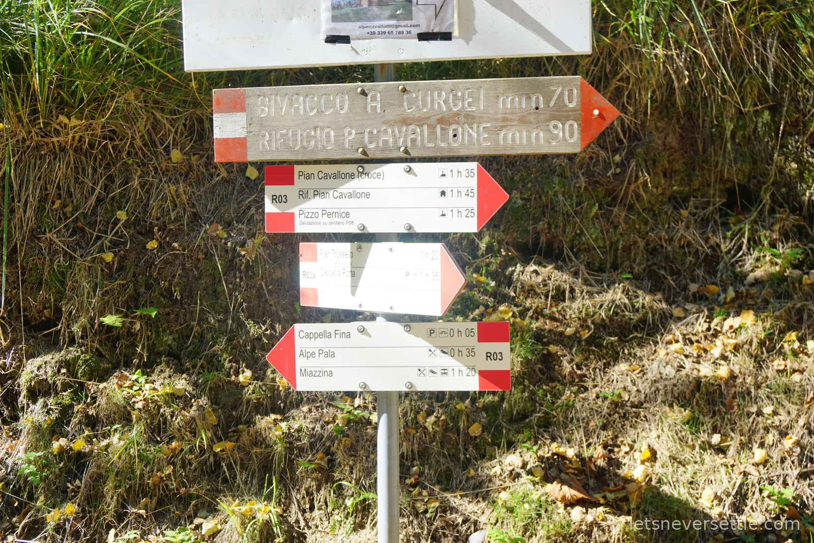A metal post on a grassy bank carrying stacked red and white walking signs reading Bivacco A. Curgei, Rifugio P. Cavallone, Pian Cavallone, Pizzo Pernice, Cappella Fina, Alpe Pala and Miazzina with walking times