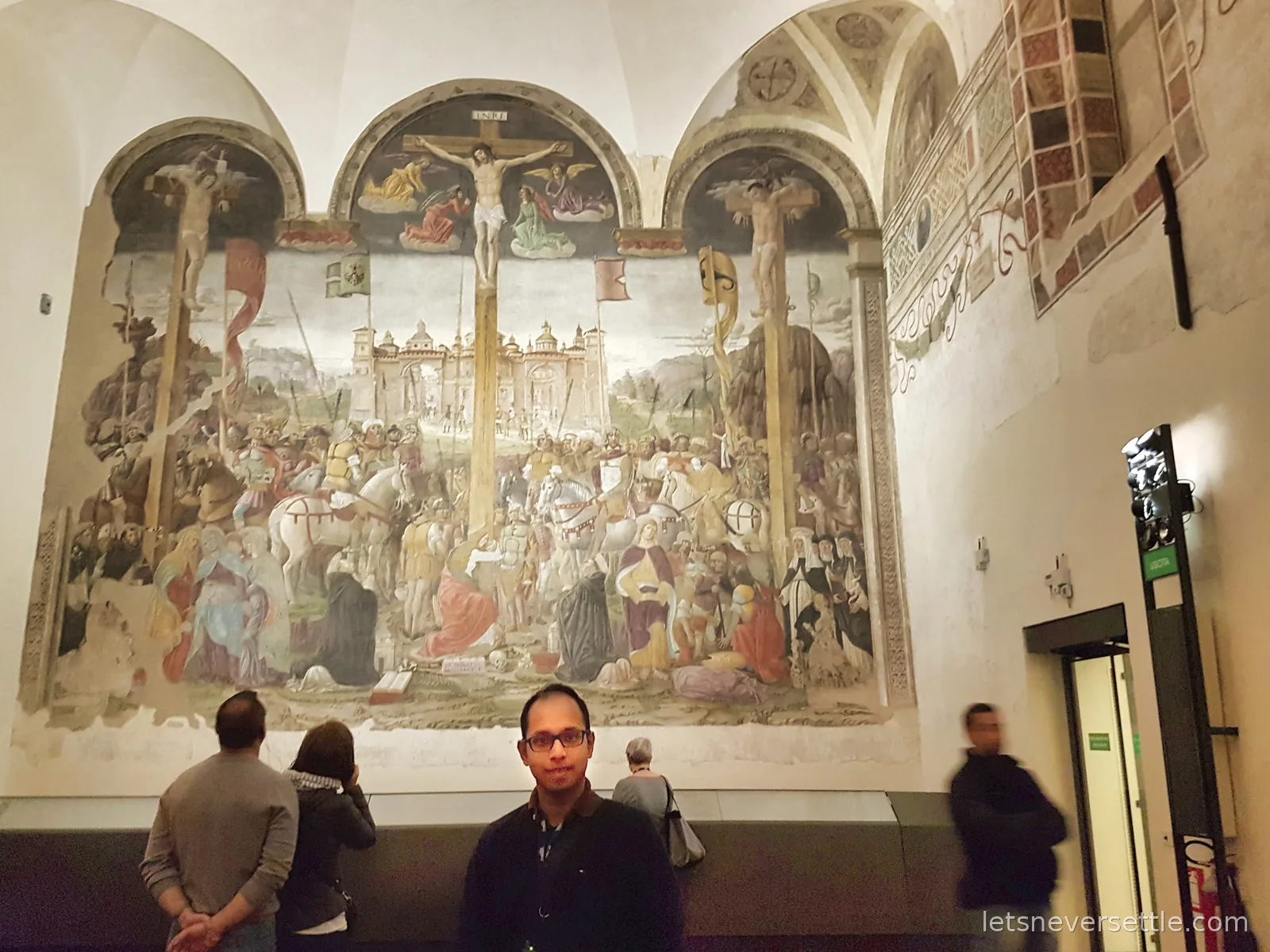 Dileep standing in front of a huge crowded Crucifixion fresco covering the opposite wall of the refectory, with visitors looking at it