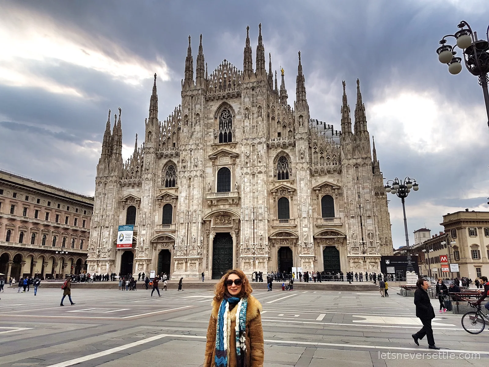 Inga standing in Piazza del Duomo in a fur collared coat with the white marble front and spires of Milan Cathedral behind her under grey cloud