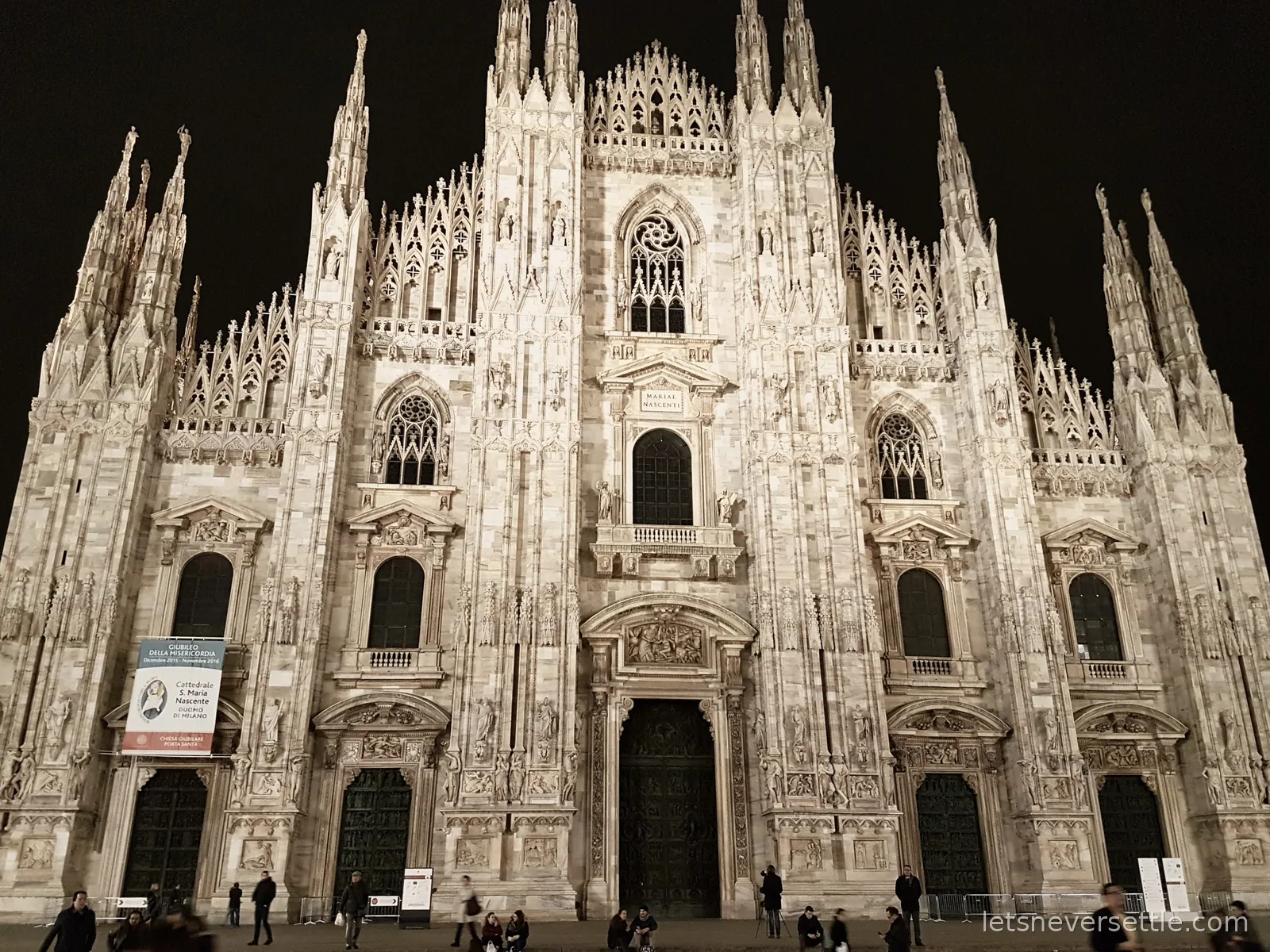 The floodlit white marble front of Milan Cathedral at night, its spires lit against a black sky, with people crossing the empty square