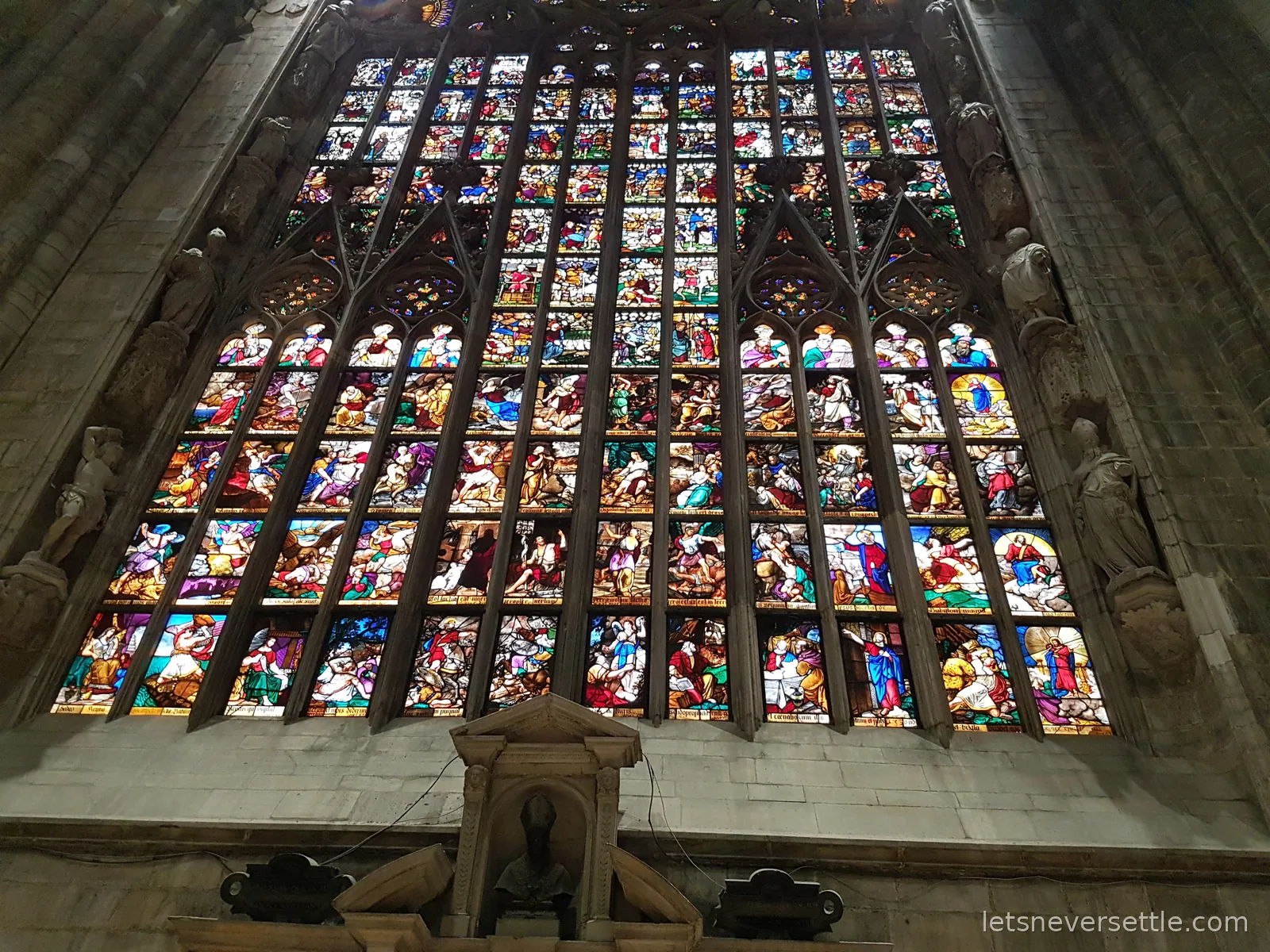 An enormous stained glass window inside Milan Cathedral, divided into hundreds of small painted panels glowing against the dark stone