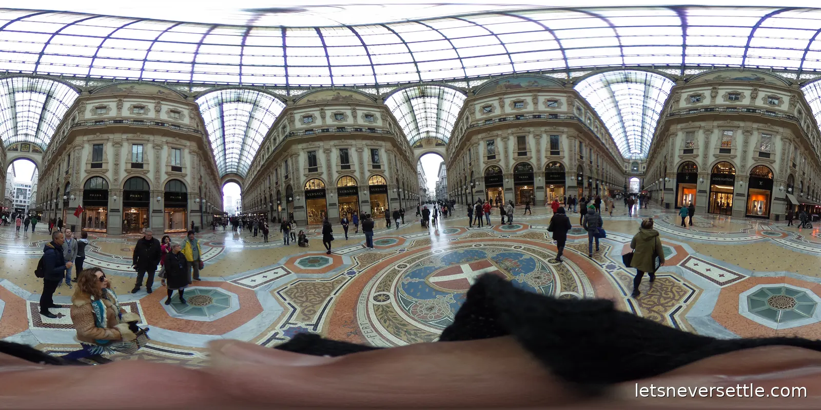 Inside the Galleria Vittorio Emanuele II, a glass roofed shopping arcade with a mosaic floor and shoppers crossing beneath the dome