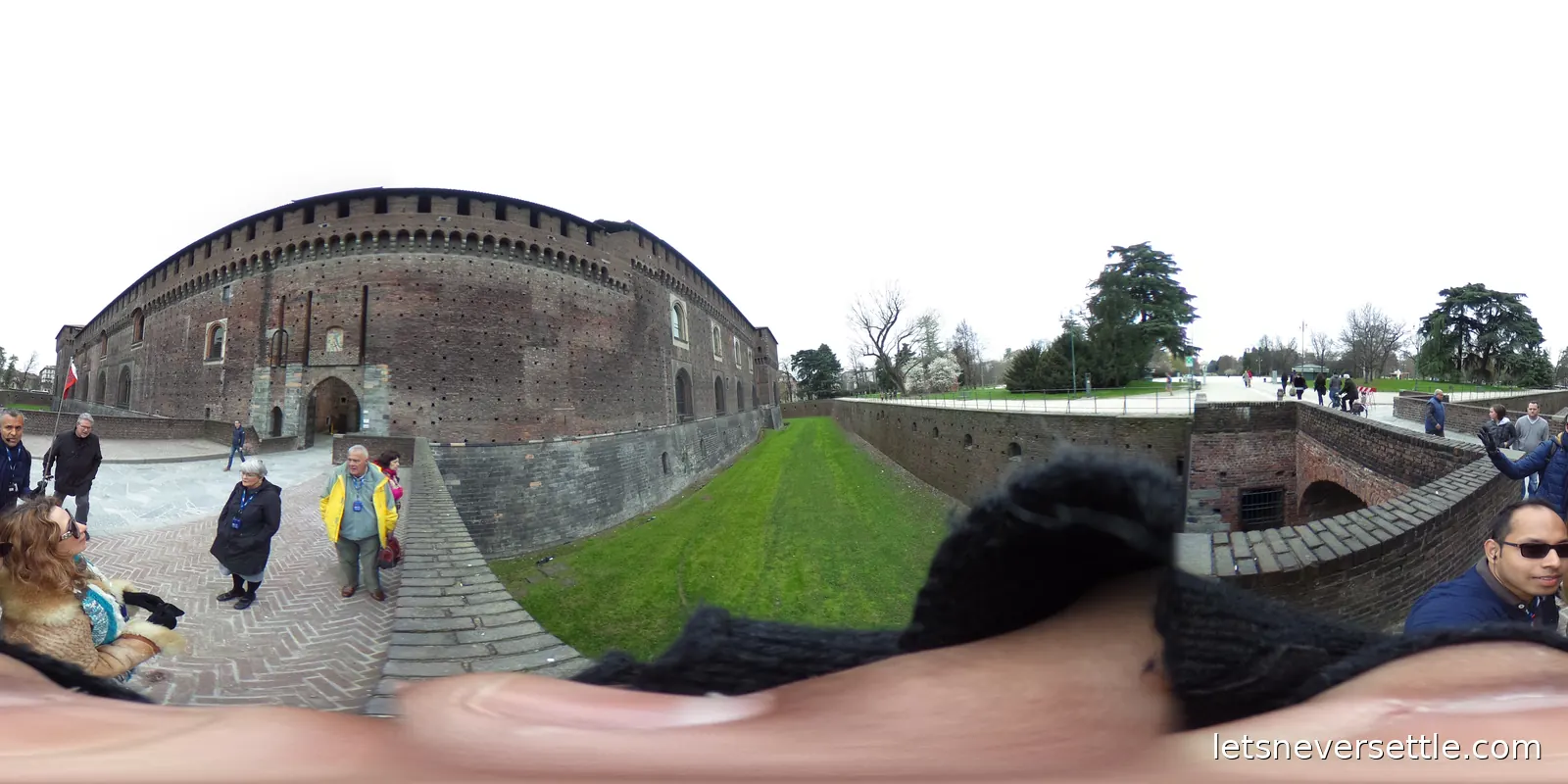 The brick outer wall and dry grass moat of Sforza Castle in Milan, with visitors walking along the path beside it