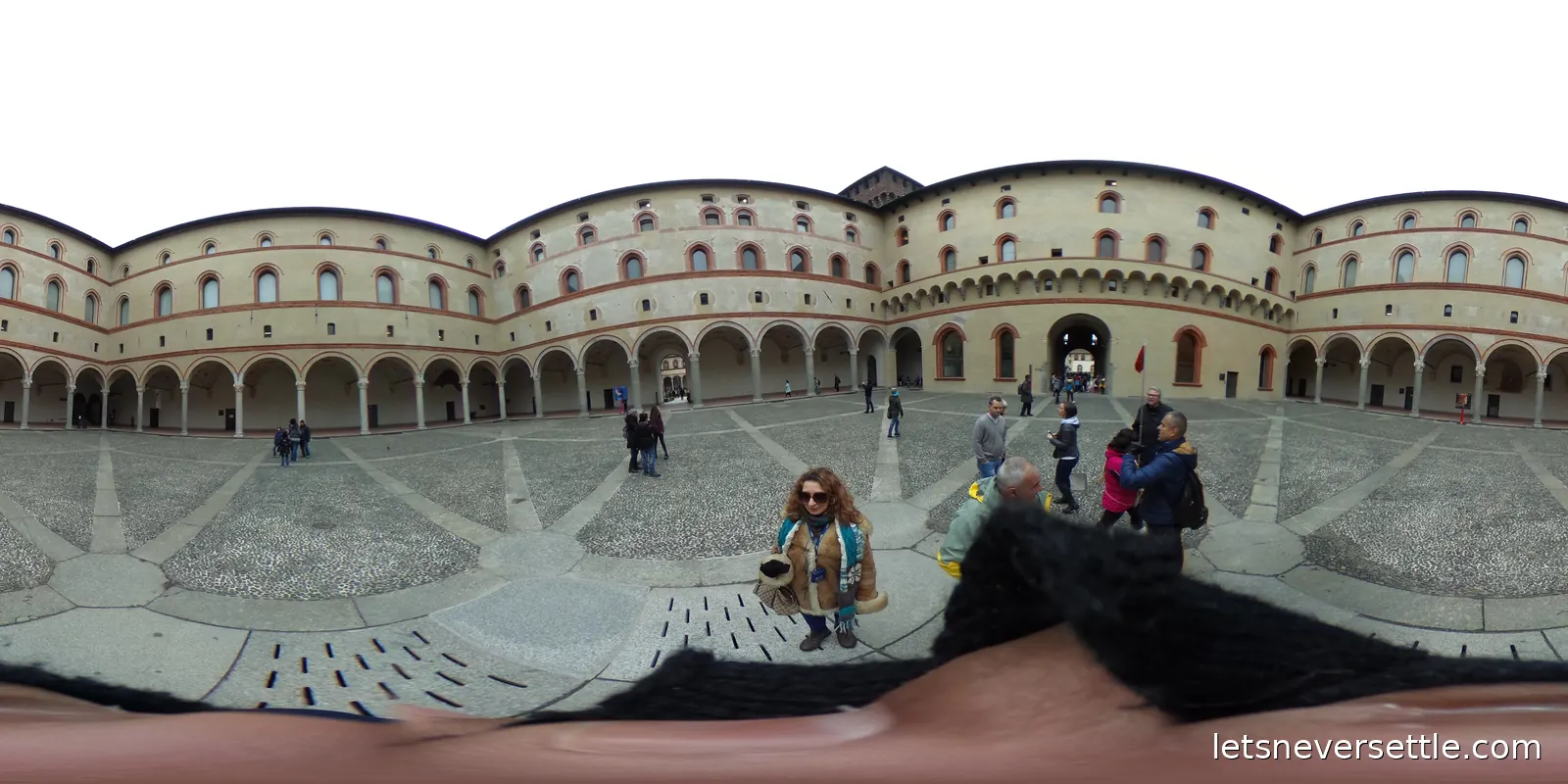 The vast inner courtyard of Sforza Castle, ringed by arcaded brick and stone ranges, with Inga standing in the middle of the gravel