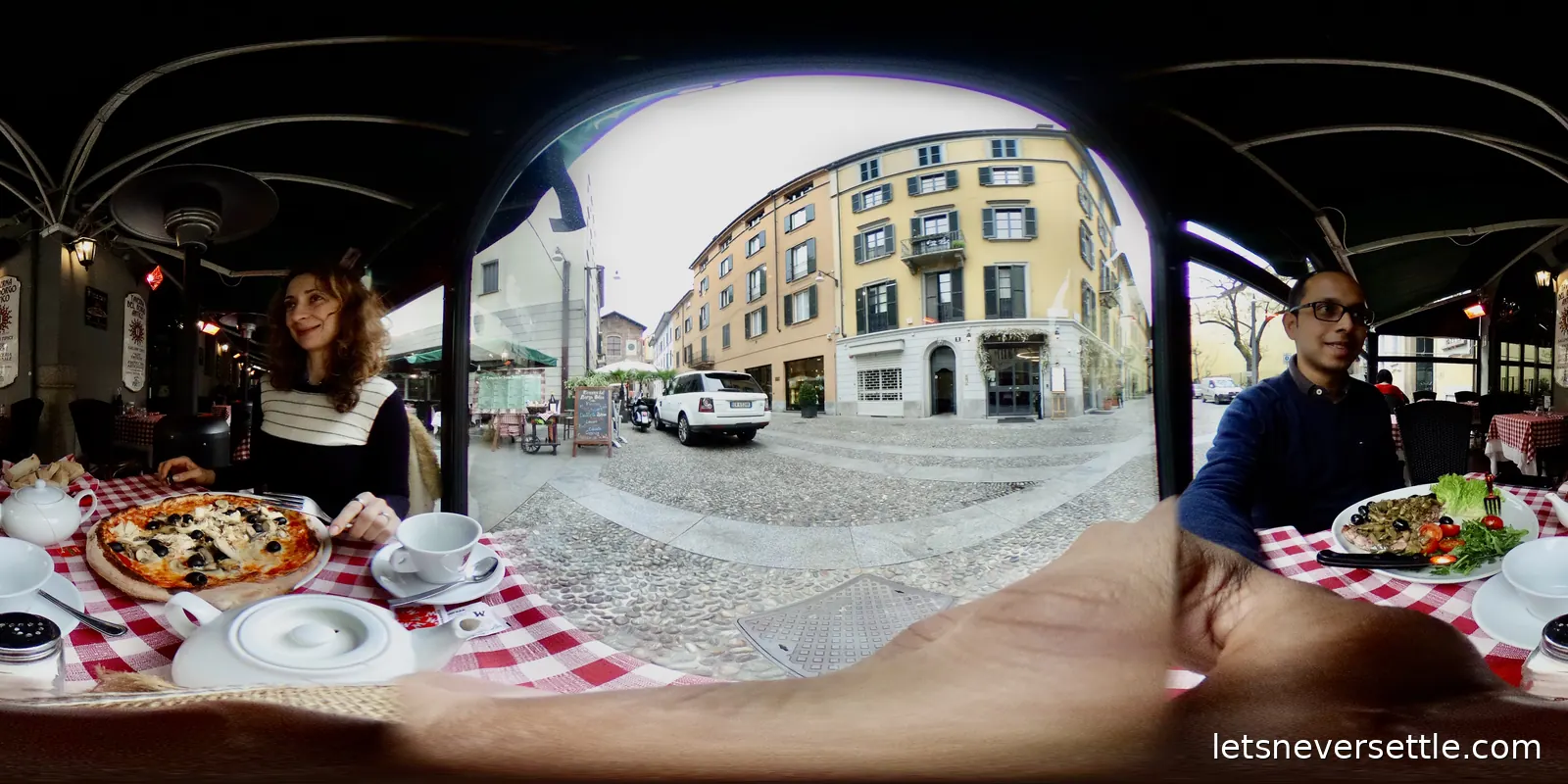 A pavement restaurant table with red checked cloth, two plates and a pizza, under an awning looking out at a Milan street corner