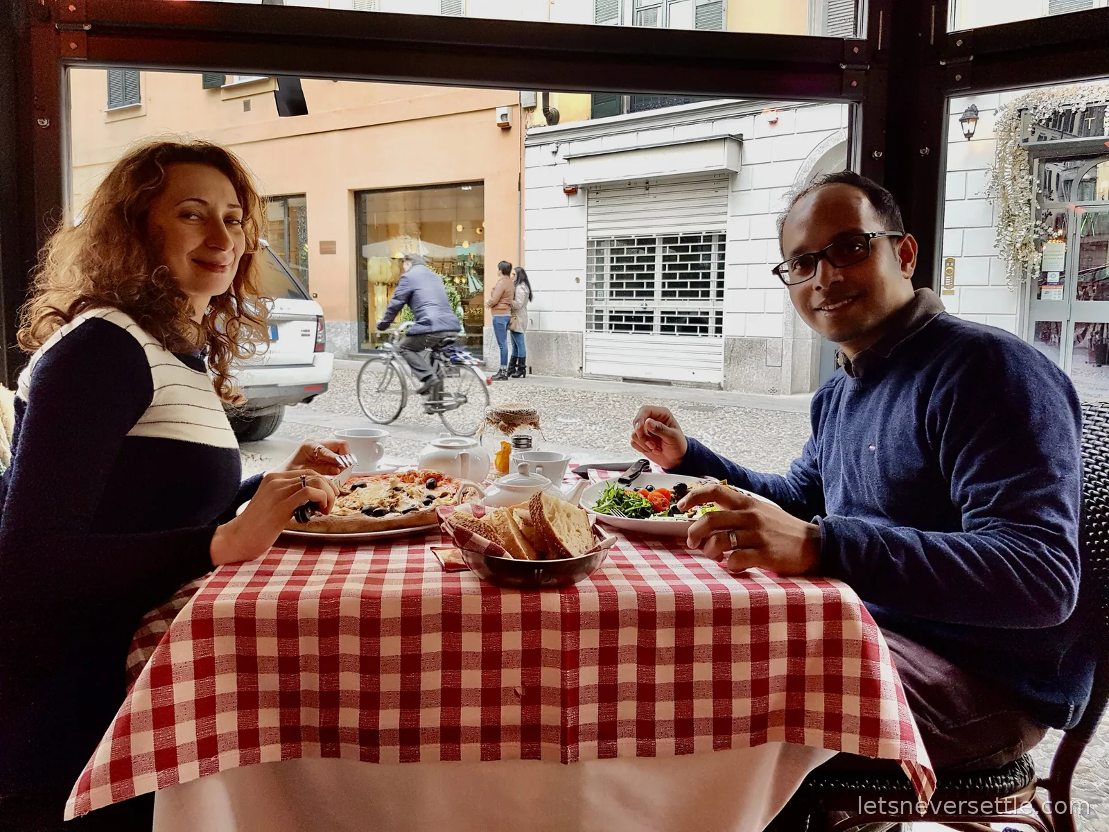 Inga and Dileep eating pizza and salad at a pavement table with a red checked cloth, a cyclist passing on the street outside