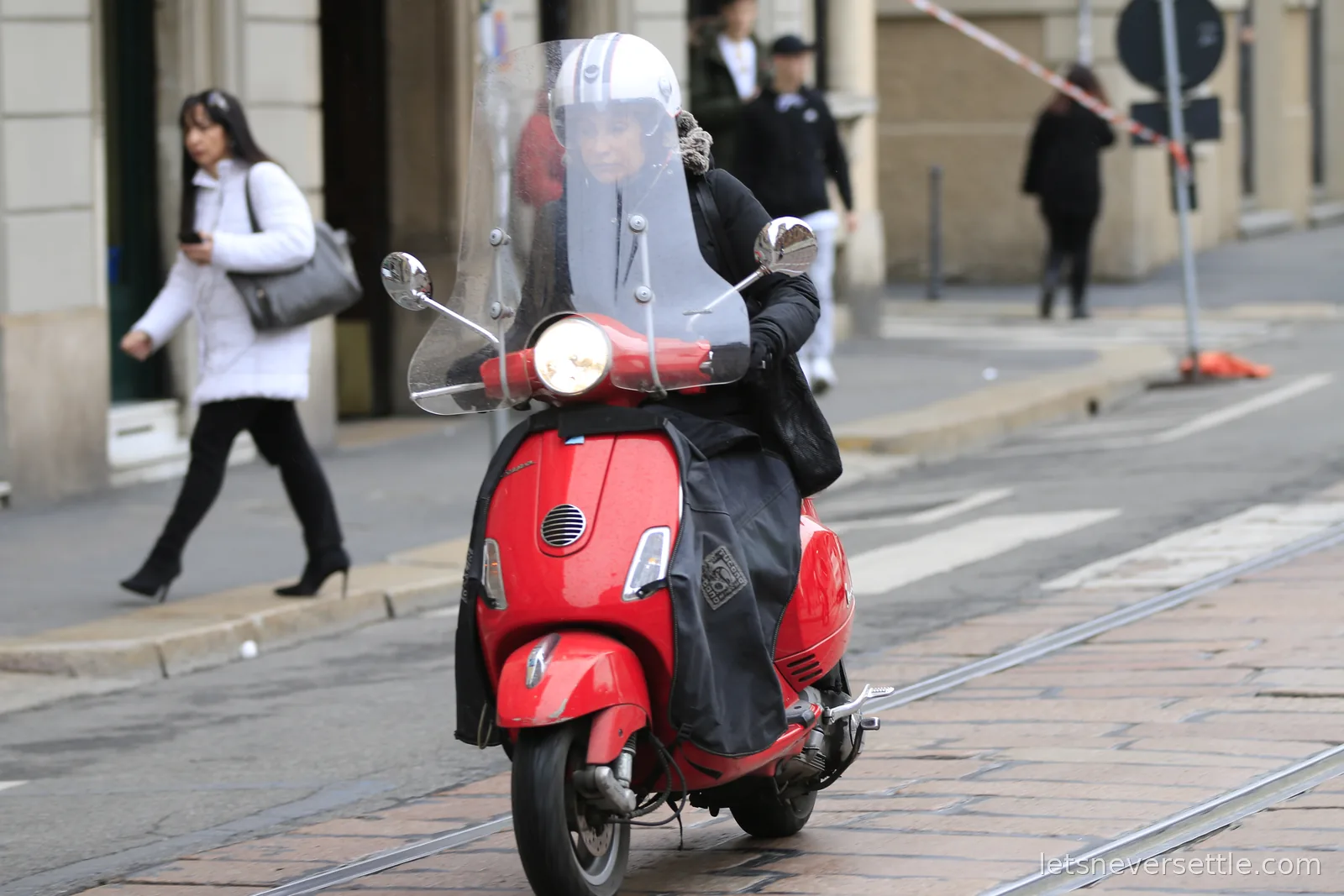 A woman in a helmet and black coat riding a red Vespa scooter with a clear windshield and a leg cover along a cobbled street with tram rails