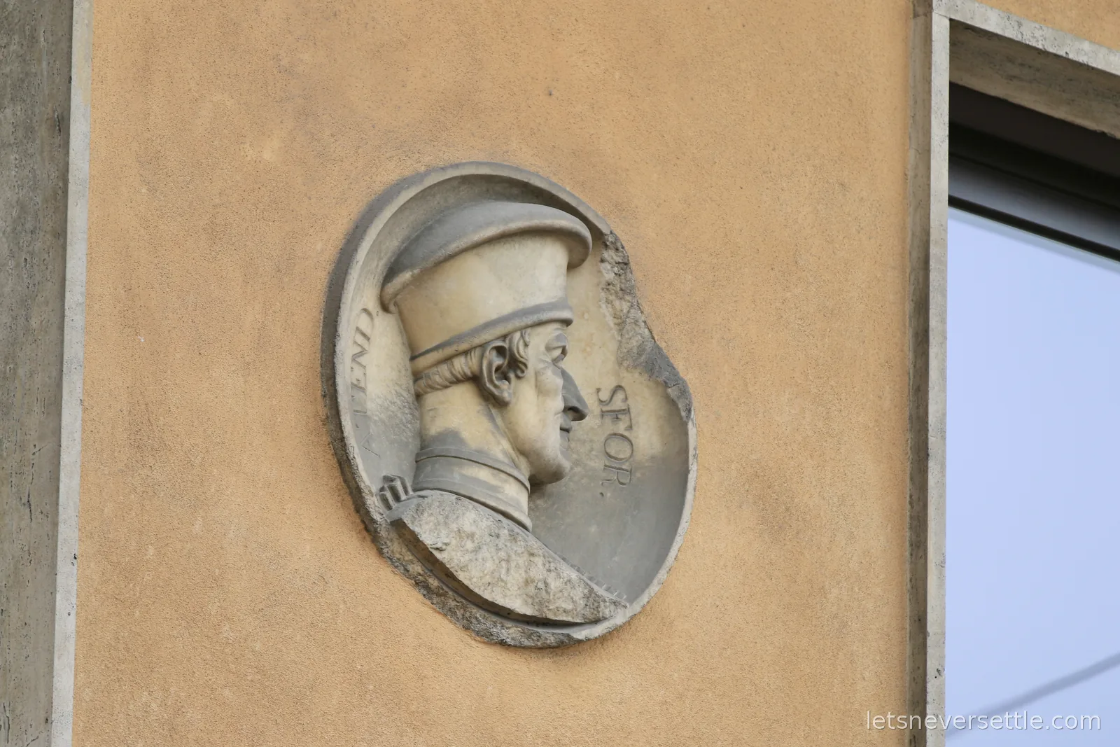 A carved stone medallion set into a plain ochre wall, showing the profile of a man in a Renaissance cap with the letters SFOR visible around the rim