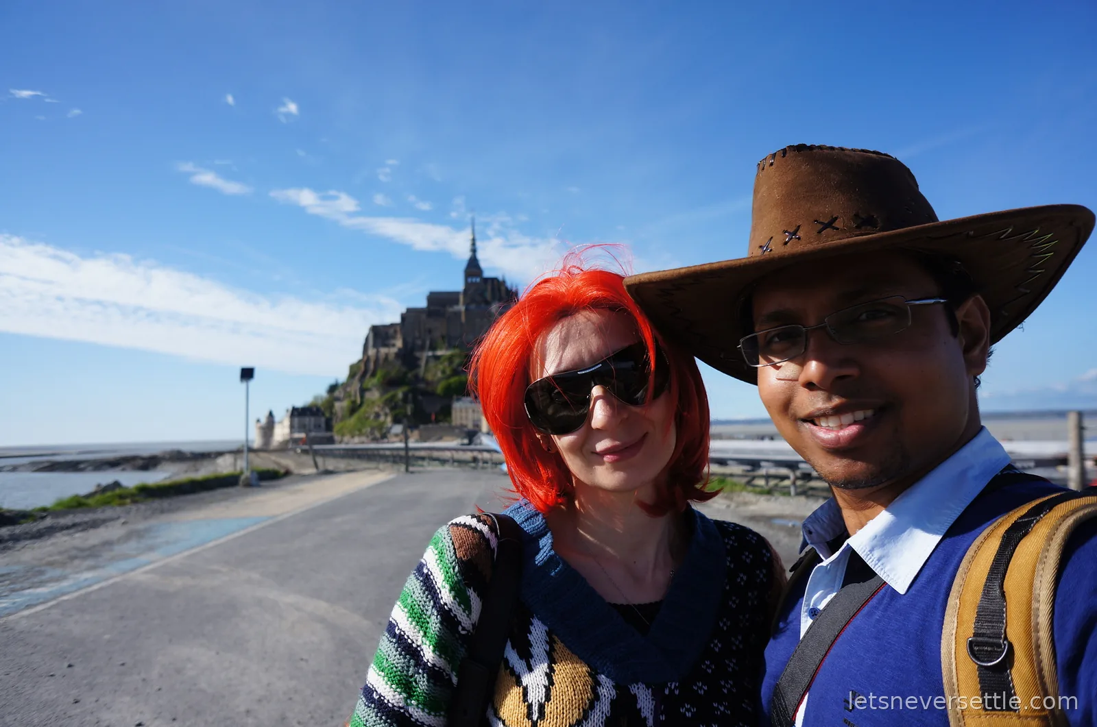 Inga and Dileep on the causeway with Mont Saint-Michel rising behind them under a blue sky
