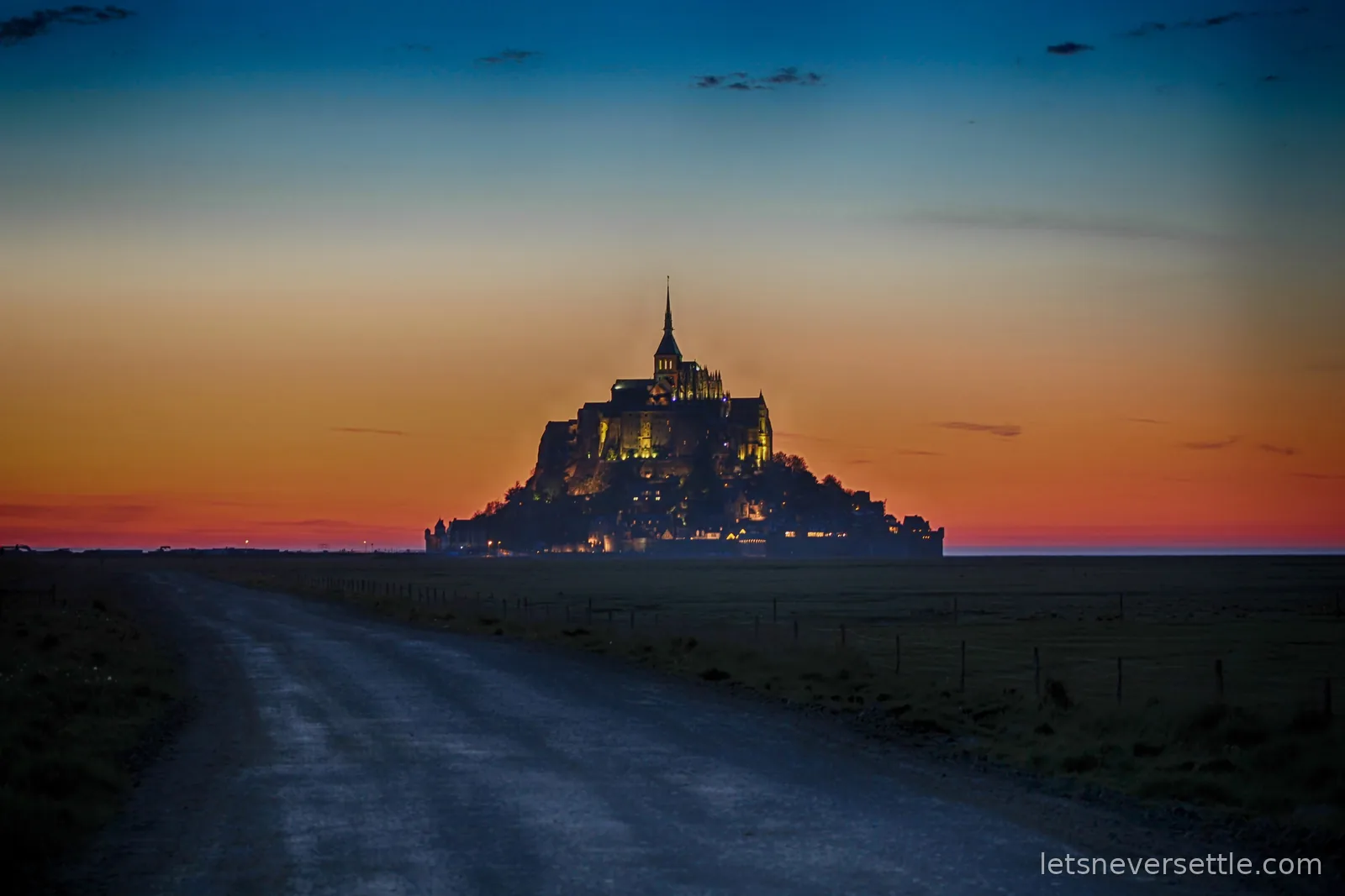 Mont Saint-Michel lit up at dusk under an orange and blue sky, seen from the dark causeway road