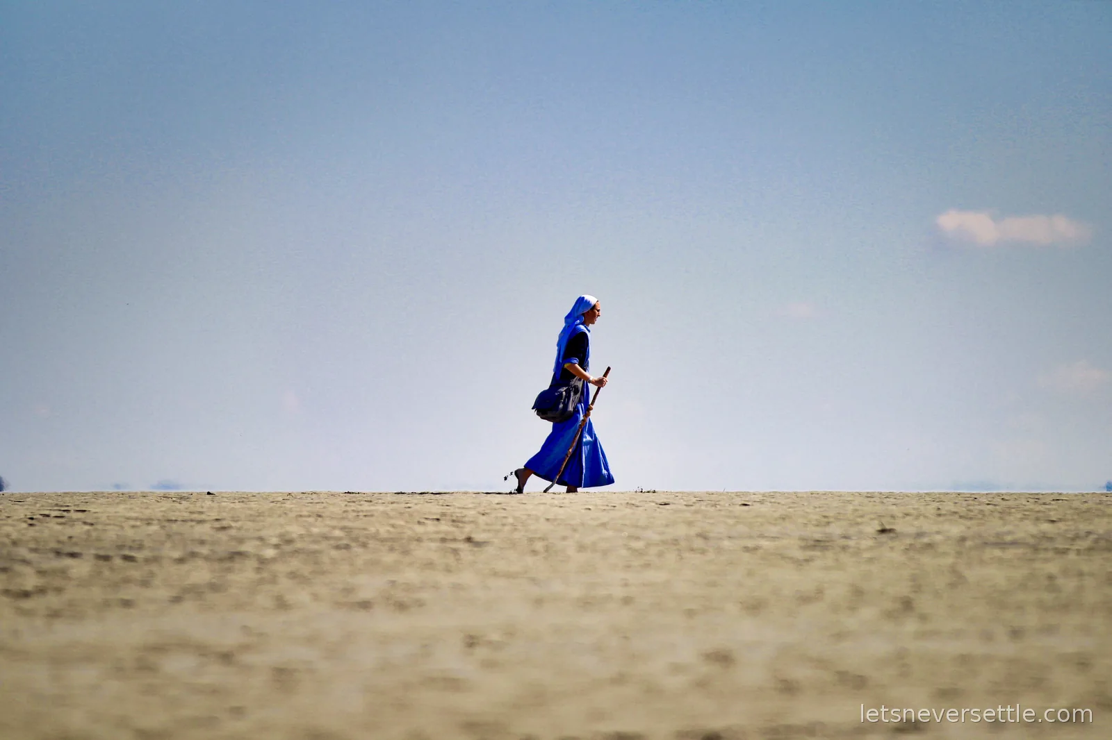 A nun in a blue habit walking barefoot across the sand flats with a wooden staff