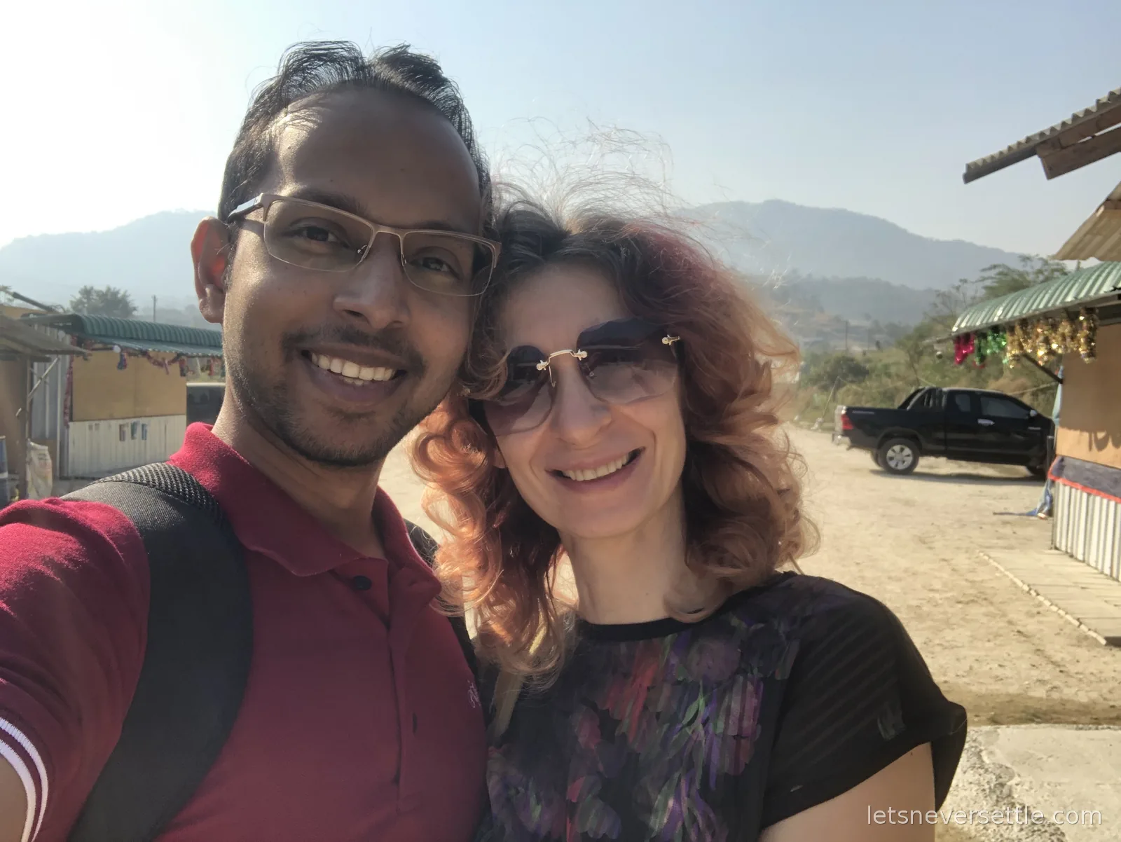 Inga and Dileep taking a selfie in bright sun at the border settlement, market stalls with gold figurines behind them