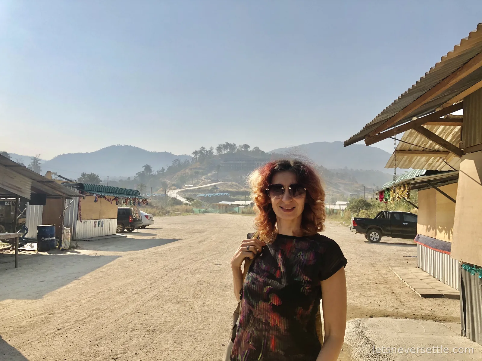 Inga standing on a dusty open lot in a Myanmar border town, corrugated stalls hung with tinsel on both sides and forested hills behind