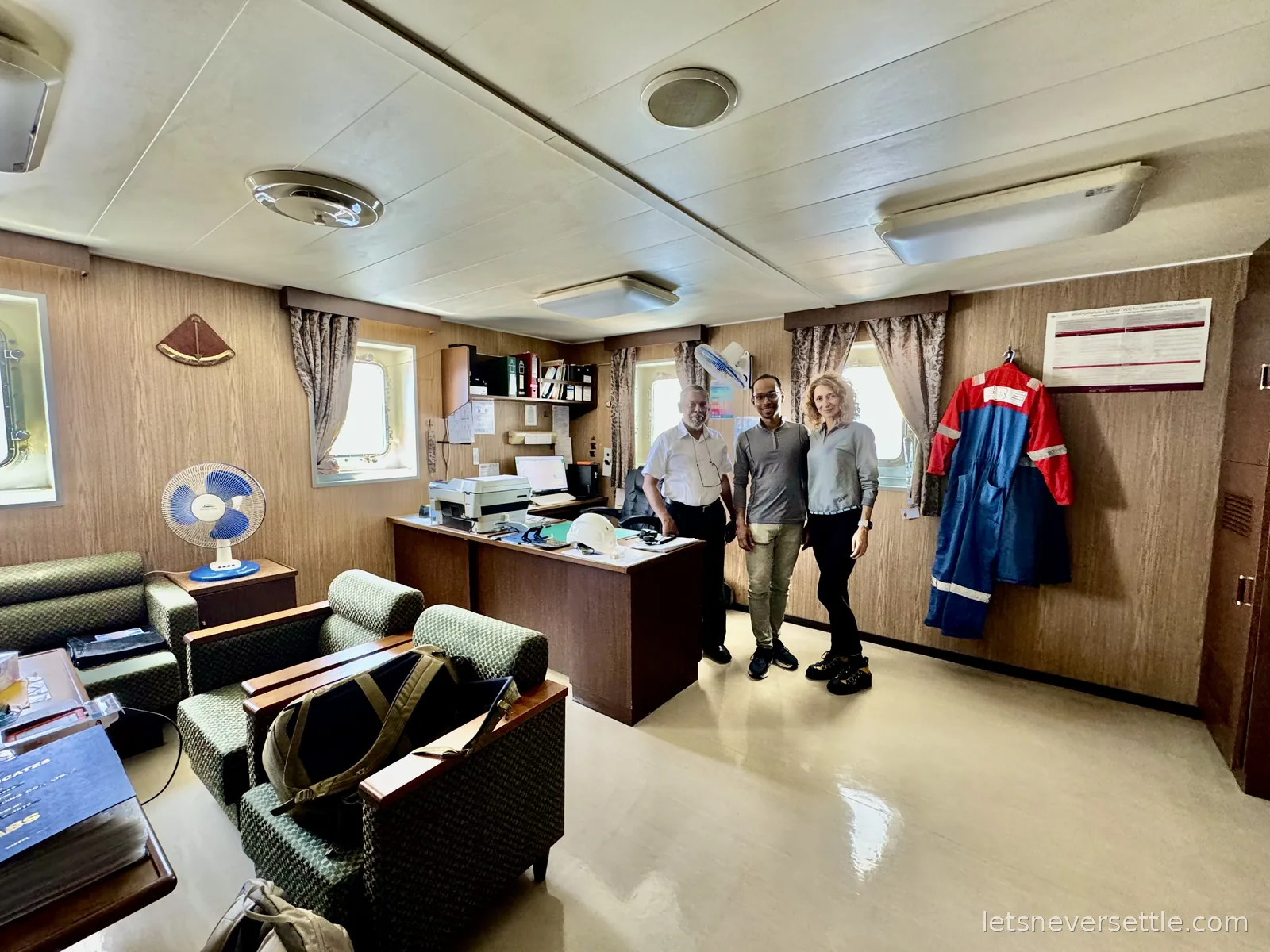 Inga and Dileep with a ship's officer in the wood-panelled day room of a cargo ship, boiler suits hanging on the wall