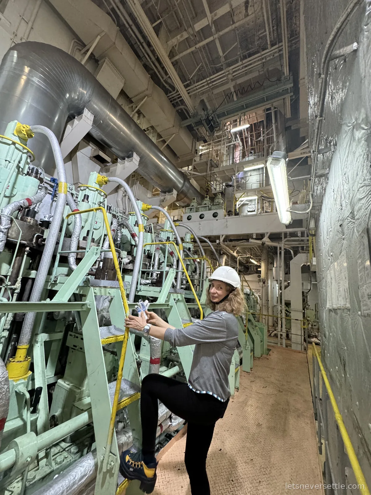 Inga in a white hard hat and hiking boots climbing on the mint green main engine block of a ship's engine room, pipes and ducting running overhead