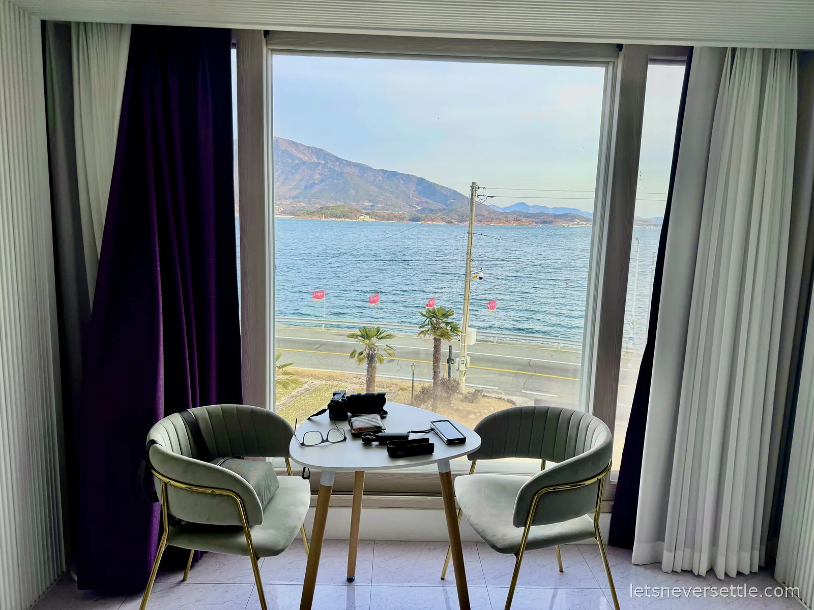 Hotel room window framing the sea, a coastal road with palm trees and mountains across the water
