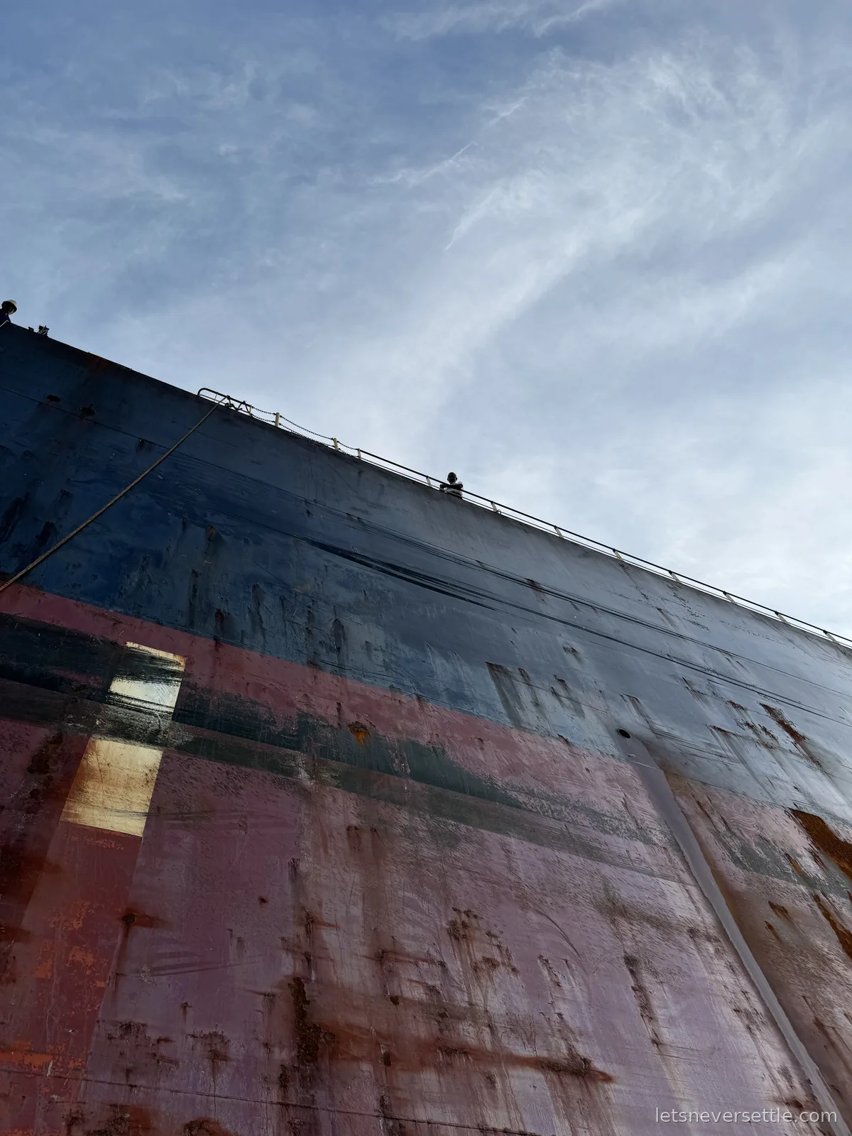 The towering rust-streaked hull of a cargo ship seen from a small boat alongside, ladder running up the side