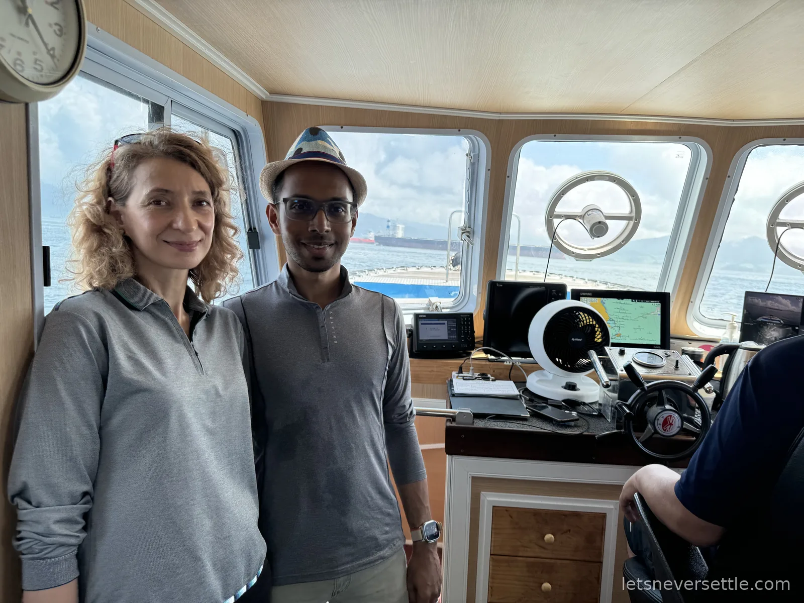 Inga and Dileep in the wheelhouse of a small boat, wheel and navigation screens beside them and the sea outside