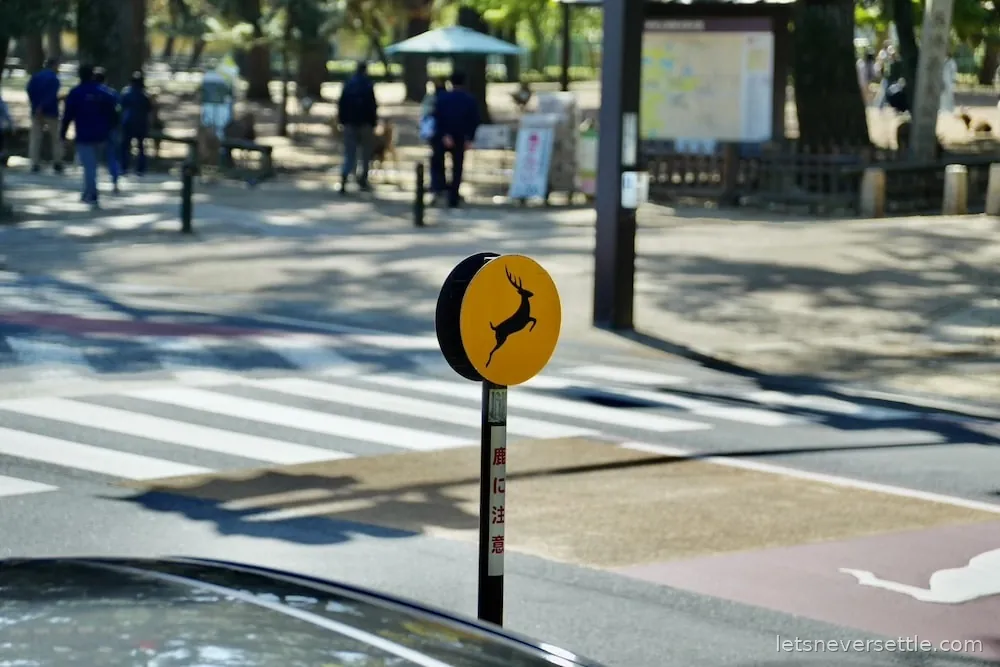 A yellow and black road sign in Nara showing a leaping deer, standing at a pedestrian crossing near the park