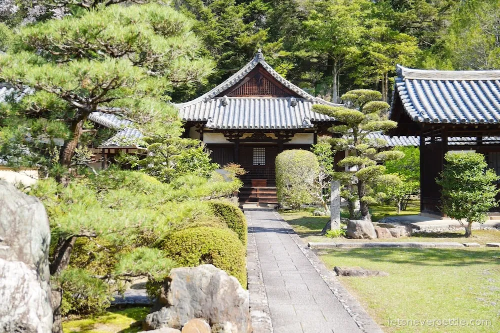 A traditional Japanese temple building in Nara with a tiled roof, behind a clipped pine and a stone-edged garden path