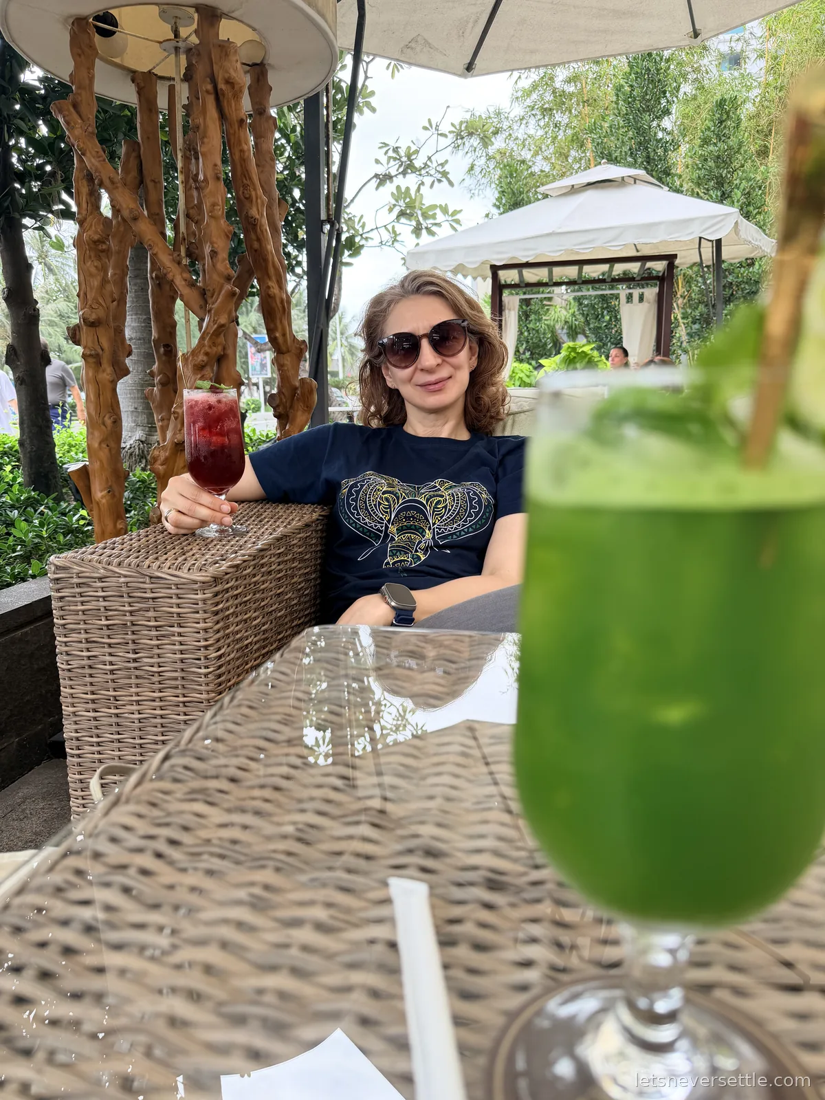 Inga in sunglasses raising a tall red fruit drink at a rattan seating area under a parasol, a bright green juice in the foreground