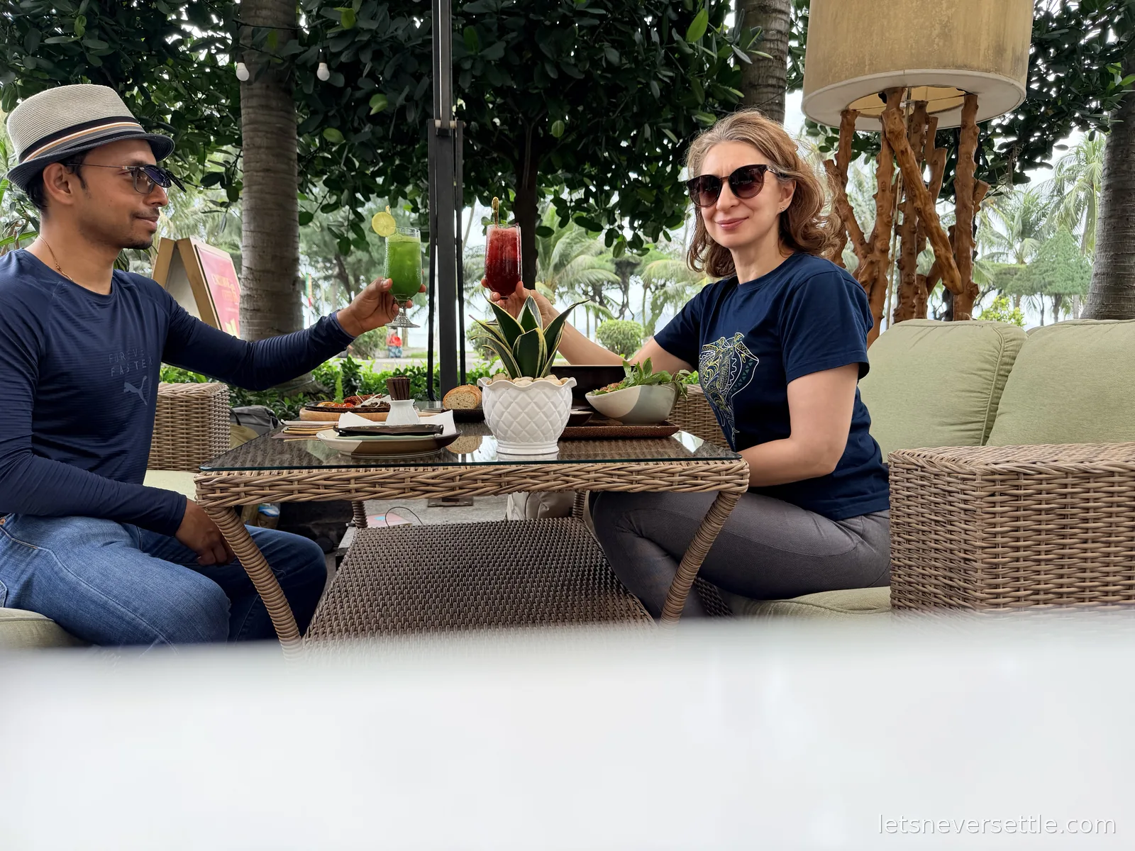Dileep in a straw trilby and Inga toasting green and red drinks across a rattan table set with small plates, palm trees behind them