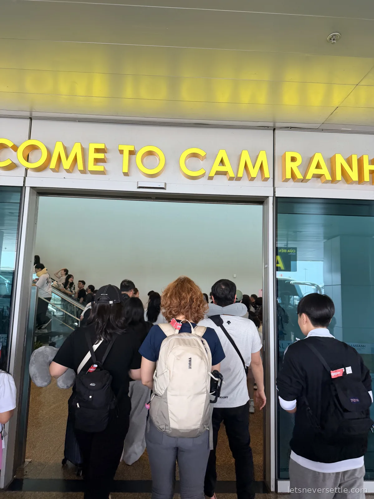 Passengers with backpacks walking through the arrivals doors at Cam Ranh airport beneath large yellow letters reading Welcome to Cam Ranh