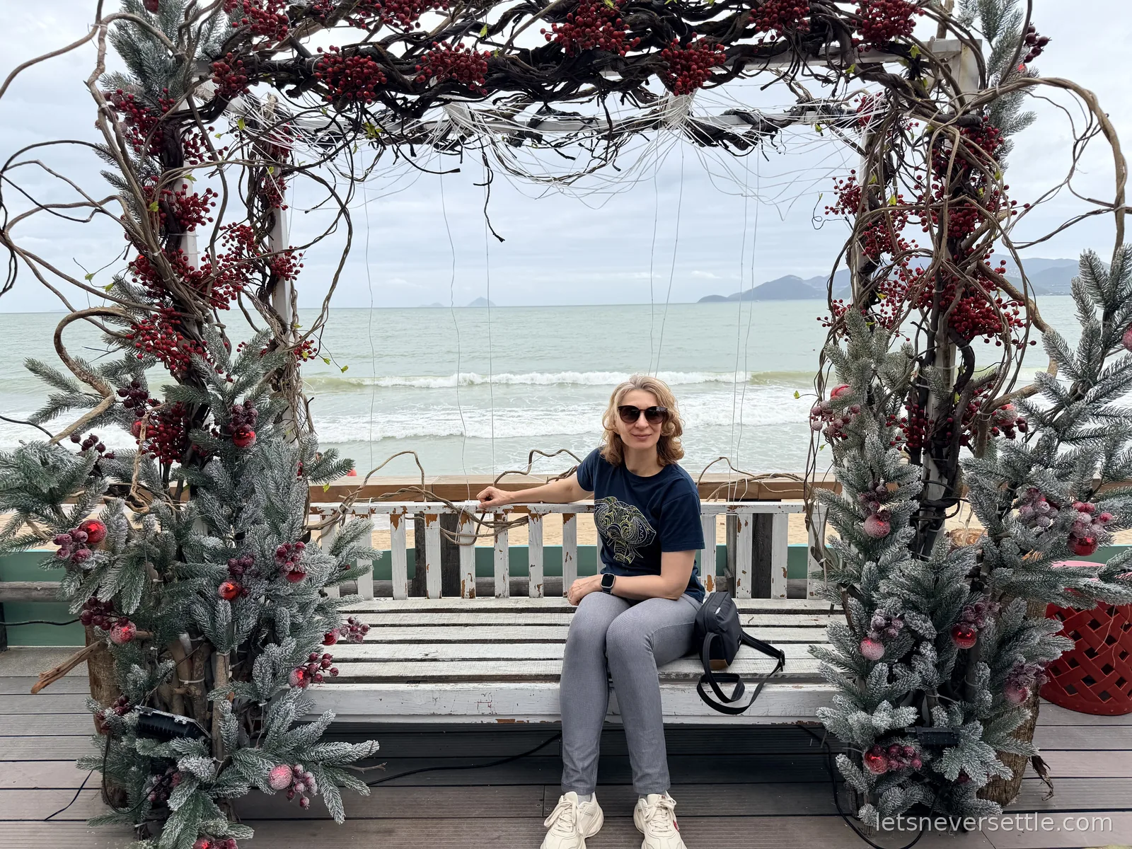 Inga sitting on a white wooden bench framed by an arch of red berries and frosted pine branches, with the grey sea and a distant headland behind