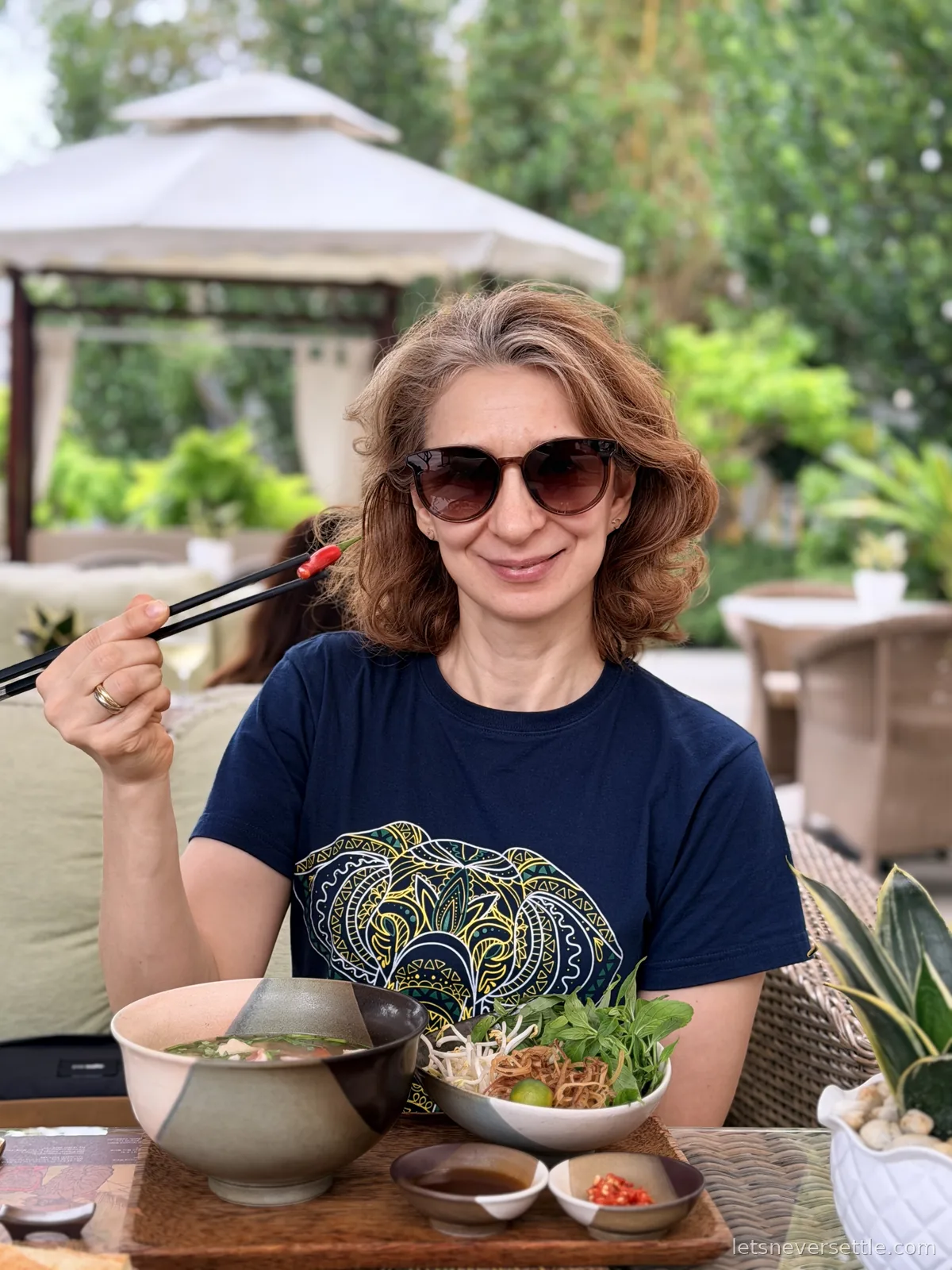 Inga in sunglasses holding chopsticks above a bowl of noodle soup, a second bowl of fresh herbs, beansprouts and lime beside it, with a garden gazebo and white parasol behind her