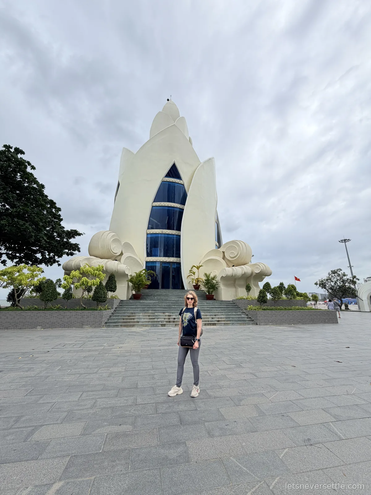 Inga standing alone on the wide empty plaza in front of the white lotus-shaped Tram Huong Tower under a flat grey sky