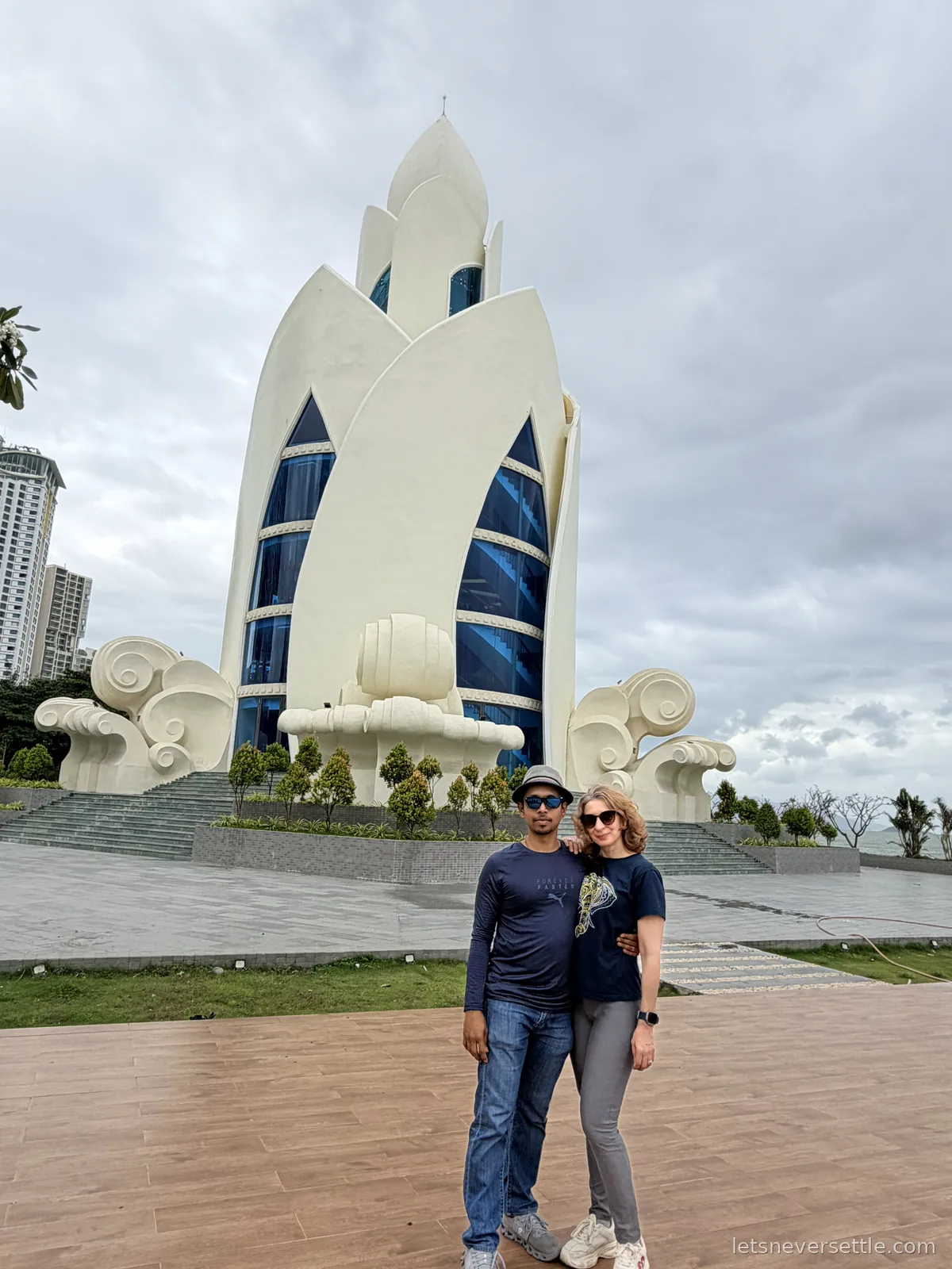 Inga and Dileep standing in front of the white lotus-bud shaped Tram Huong Tower on Nha Trang's seafront square under heavy grey clouds