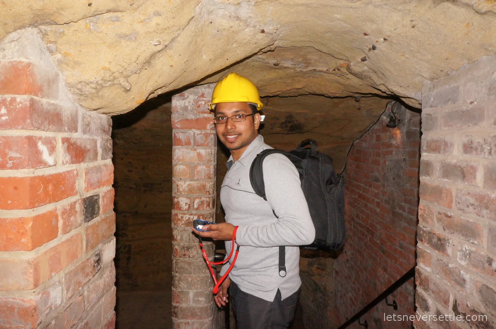 Dileep in a yellow hard hat and backpack standing among brick pillars and carved sandstone in a cave beneath Nottingham