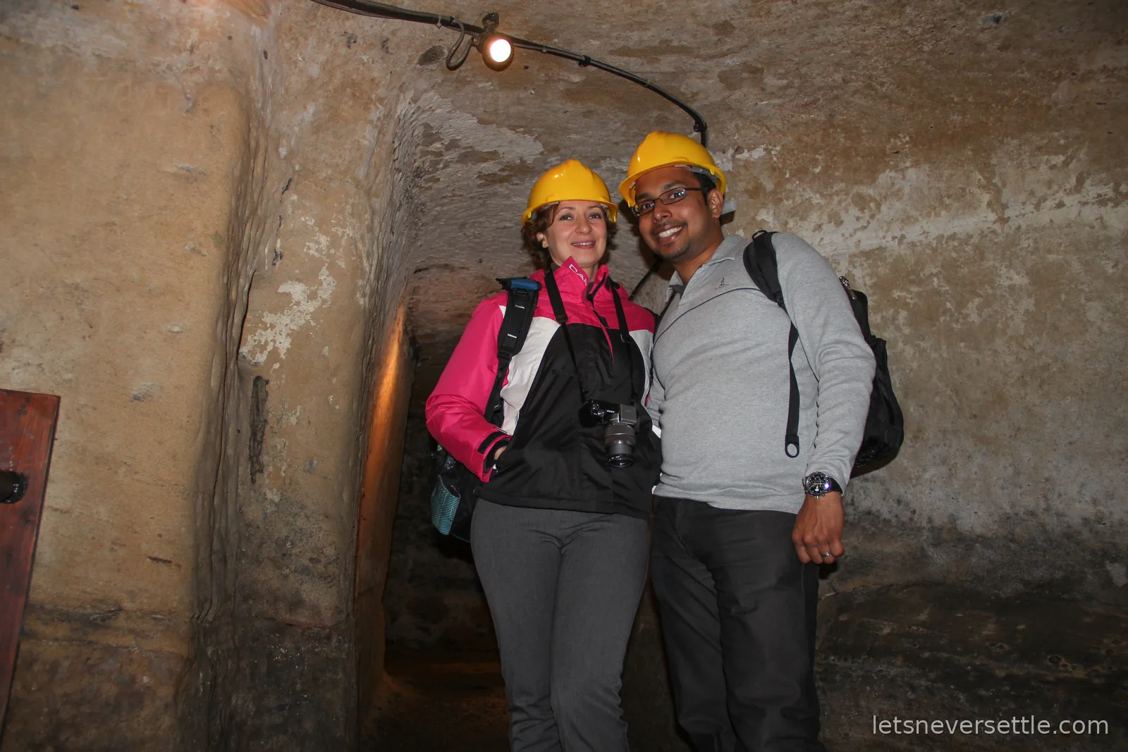 Inga and Dileep smiling in yellow hard hats in a low arched sandstone passage lit by a single lamp