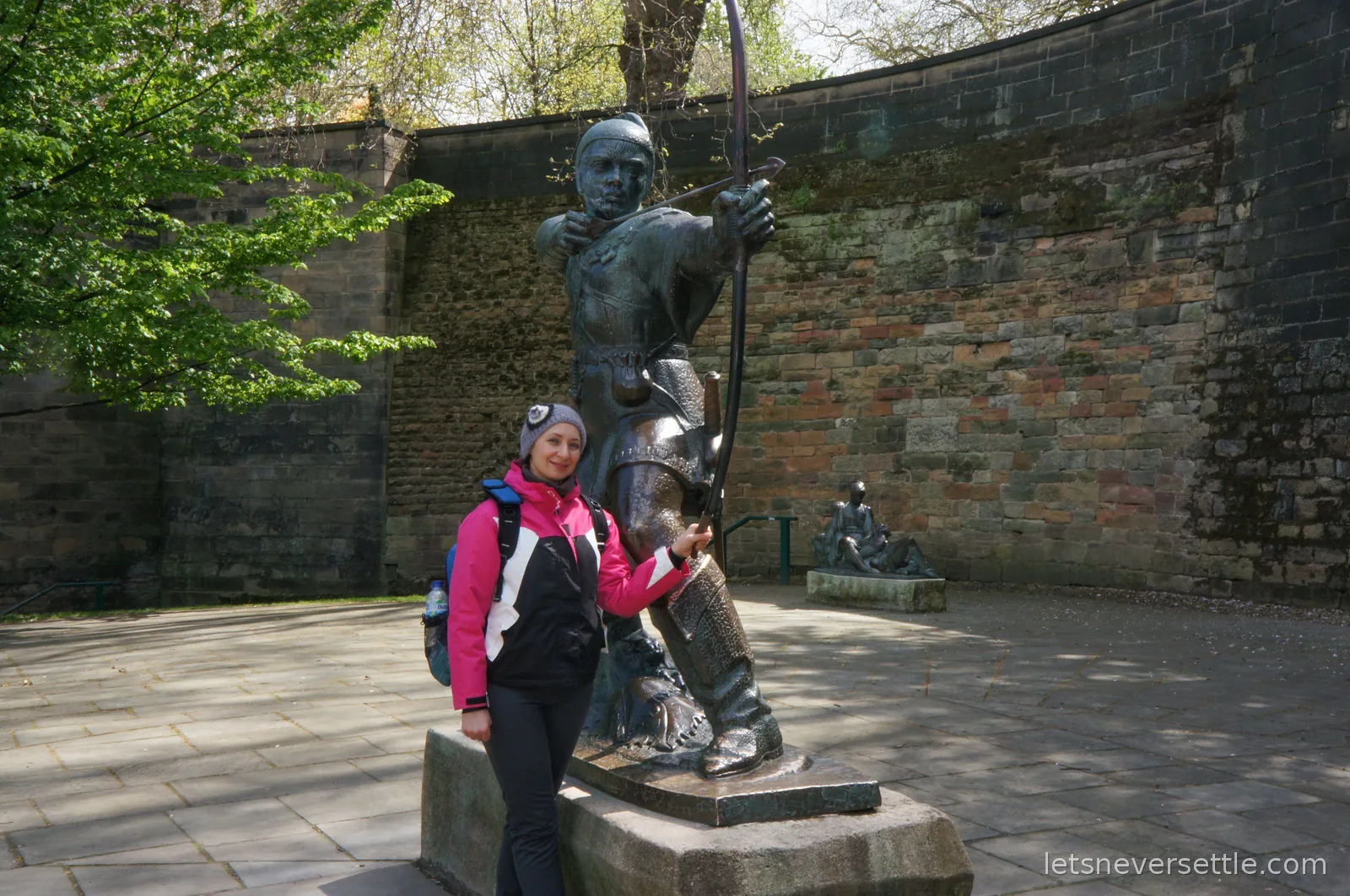 Inga in a pink and black jacket standing with the bronze Robin Hood statue drawing his bow, the stone wall of Nottingham Castle curving behind