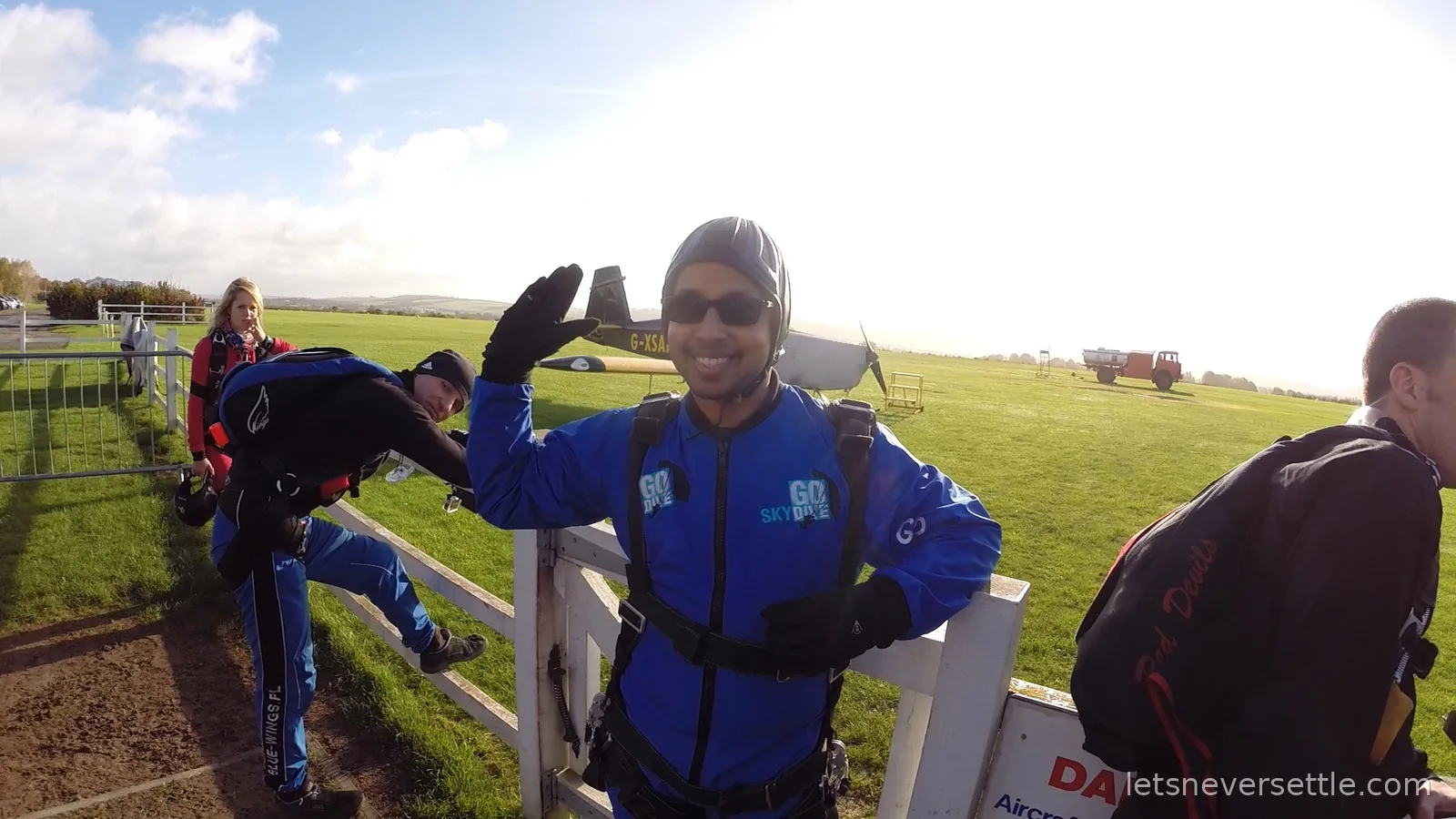 Dileep in a blue GoSkydive jumpsuit waving beside a white gate on the green airfield with a light aircraft and instructors behind