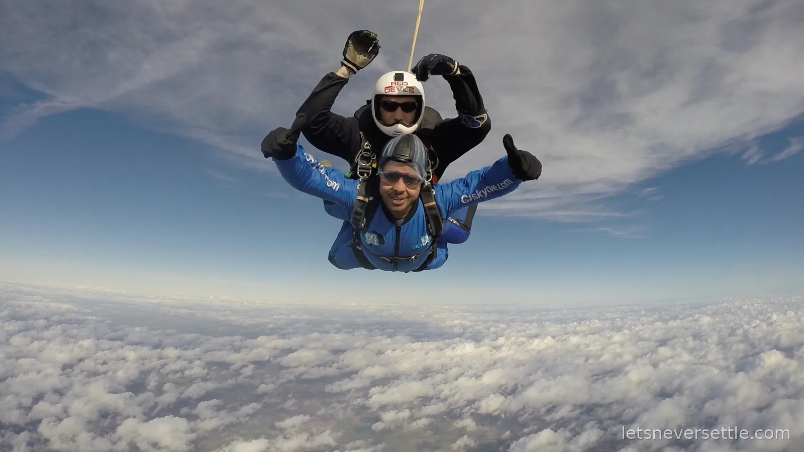 Dileep in a blue GoSkydive jumpsuit in freefall with his tandem instructor, thumbs up above a floor of cloud