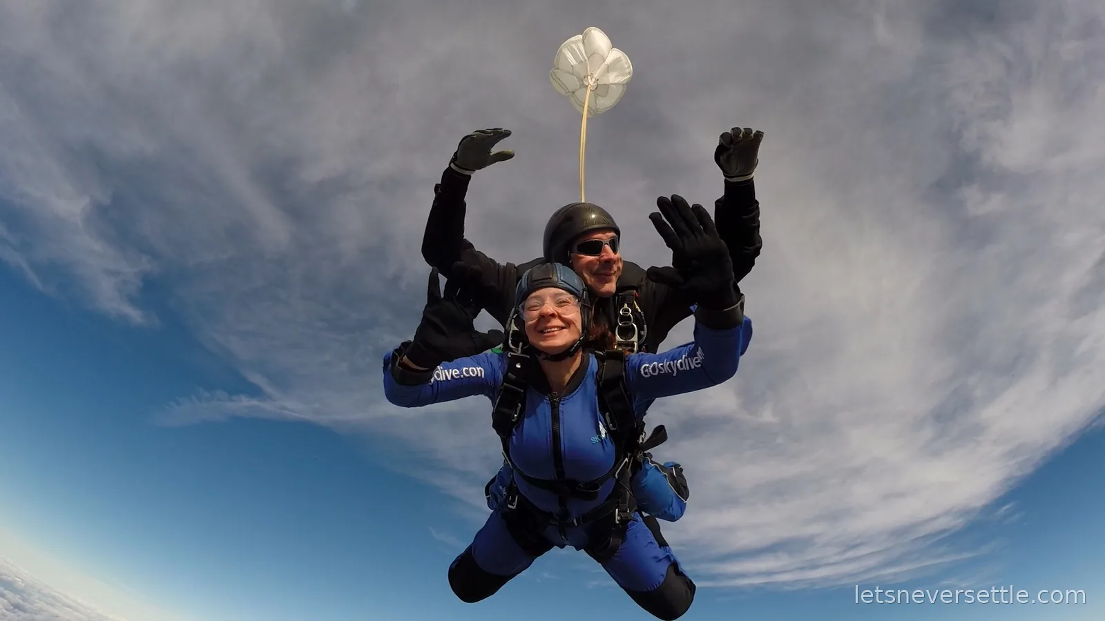 Inga in freefall strapped to her tandem instructor with arms raised, the drogue chute trailing above against a swirl of cloud
