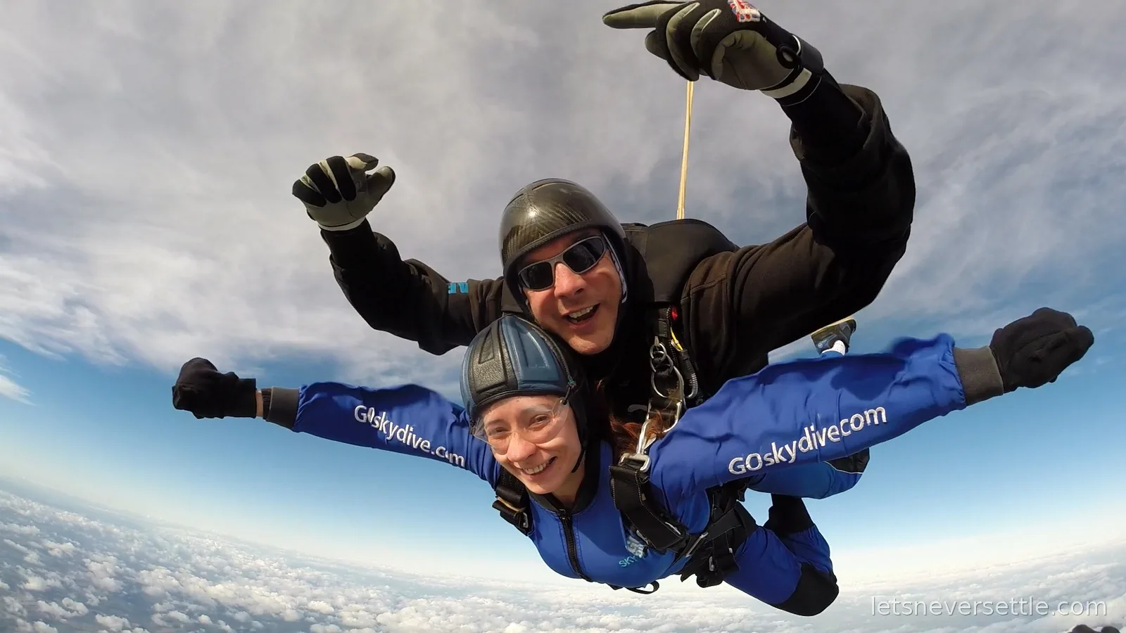 Inga grinning in freefall with her tandem instructor above a floor of broken cloud and distant fields