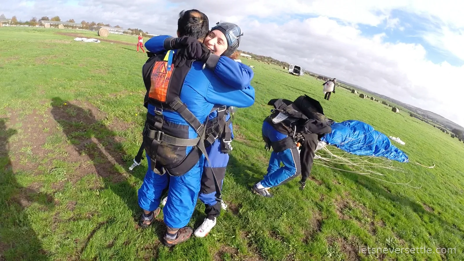 Inga hugging her tandem instructor on the landing field with a blue parachute bundled on the grass behind