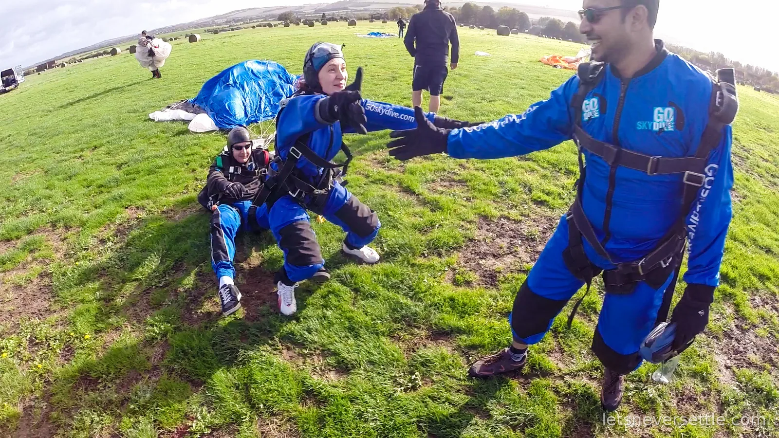 Inga kneeling on the grass giving a thumbs up after landing, parachutes and hay bales scattered across the field, Dileep laughing at the edge of the frame
