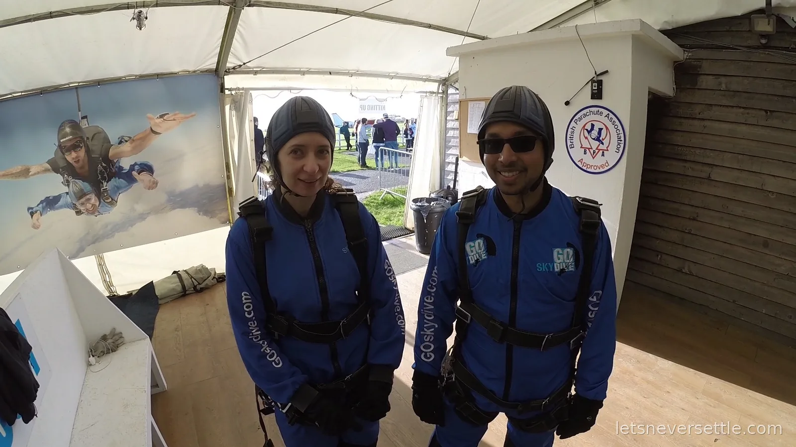 Inga and Dileep in blue GoSkydive jumpsuits and soft helmets standing in the kit tent beside a poster of tandem skydivers