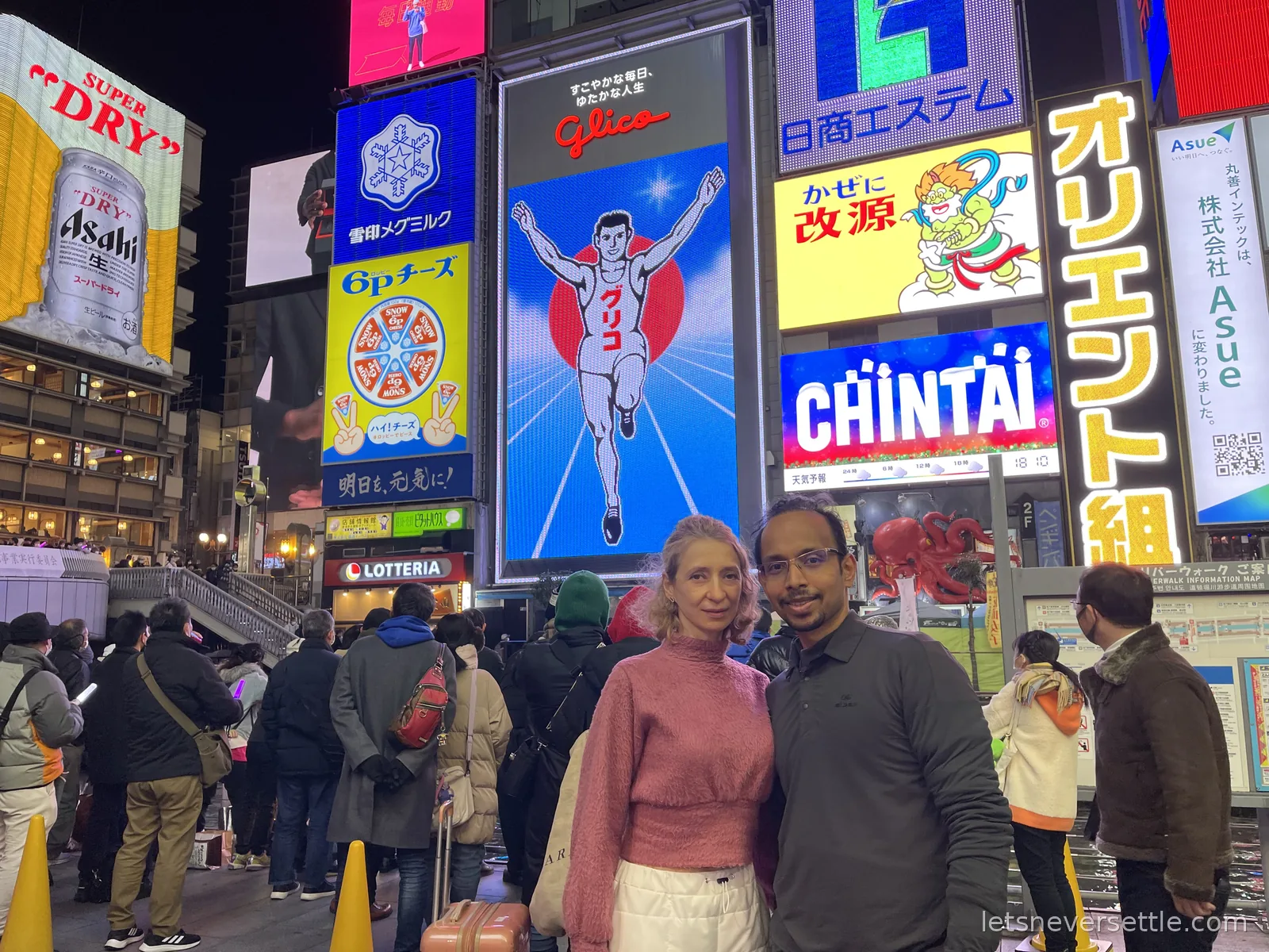 Inga and Dileep at night in Dotonbori in front of the illuminated Glico running man billboard and a wall of neon signs