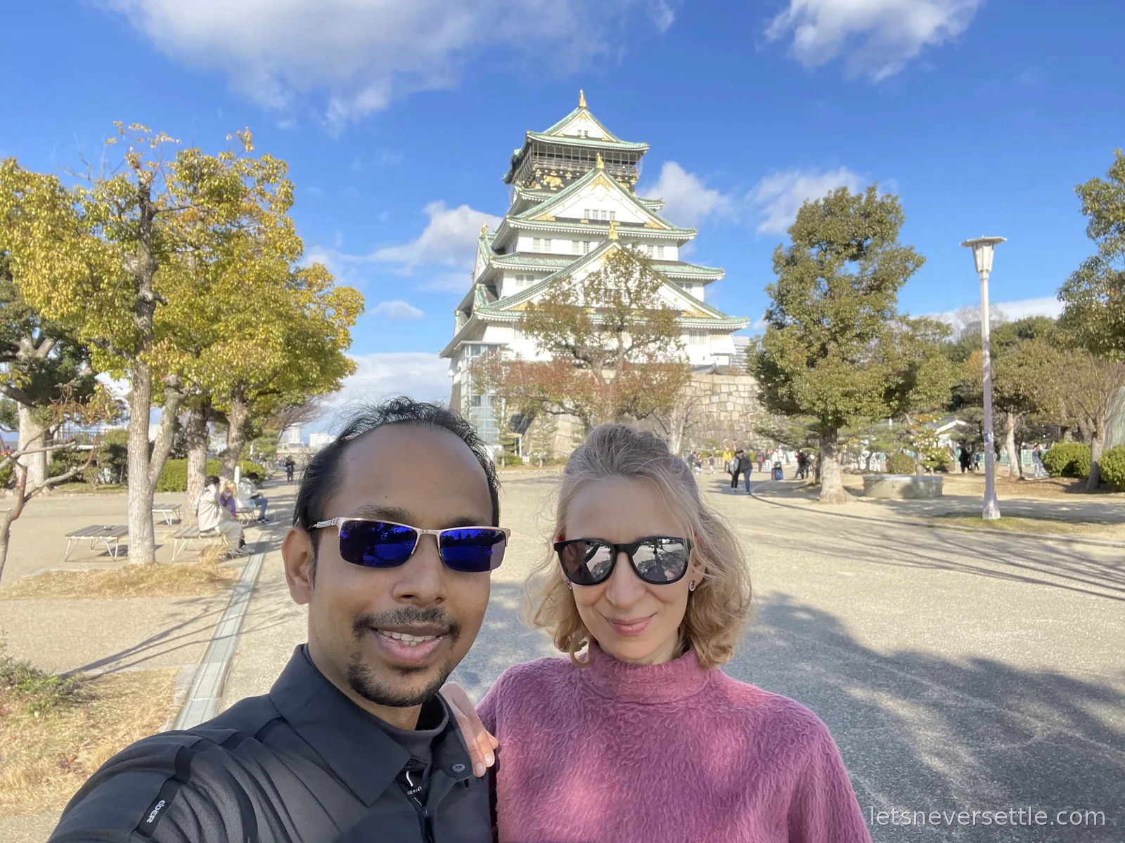 Selfie of Dileep and Inga in front of Osaka Castle, its green roofs and gold trim rising above the trees under a blue sky