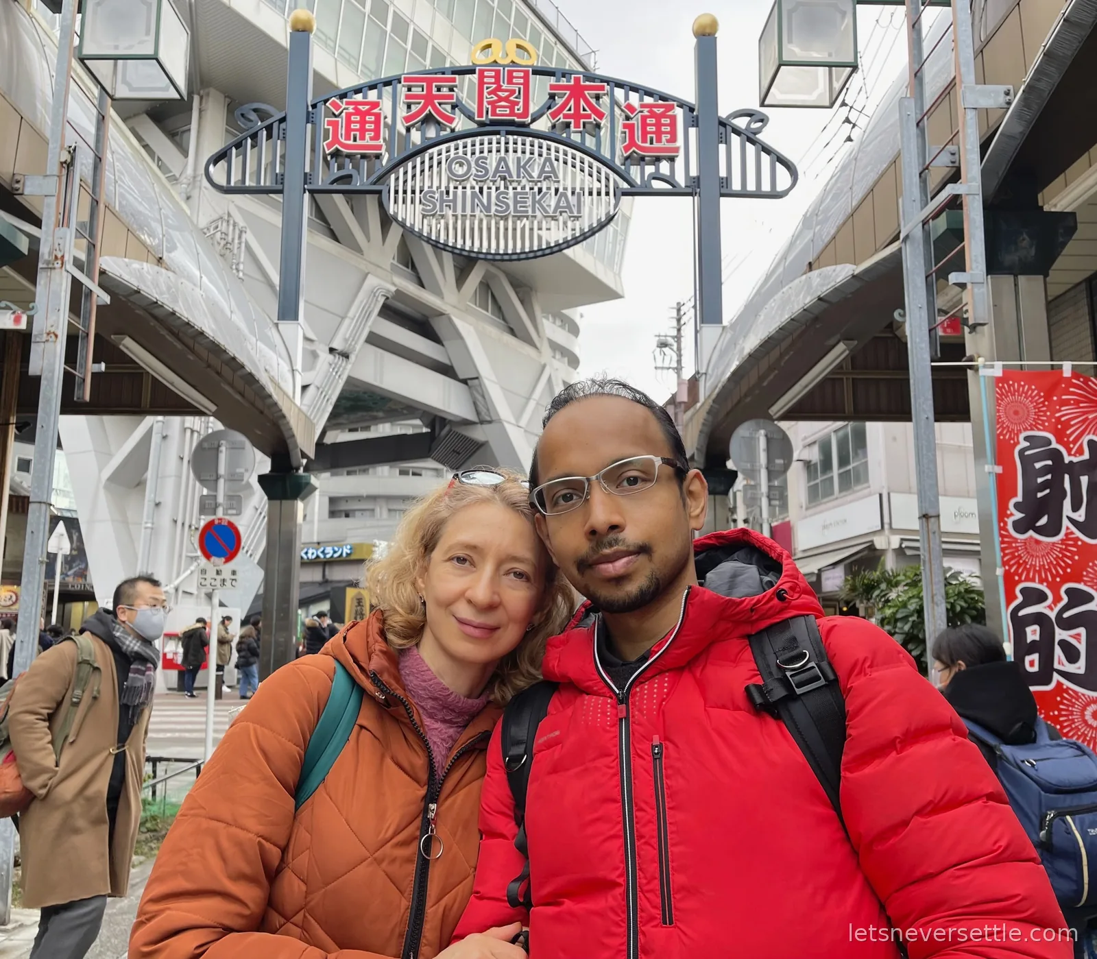 Dileep and Inga in winter jackets under the Osaka Shinsekai entrance arch with Tsutenkaku Tower rising behind it