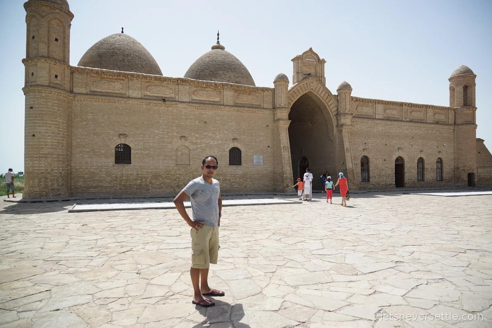 Dileep standing on the flagstone forecourt of the Arystan Bab Mausoleum, a long brick building with two domes, corner towers and a pointed arch portal