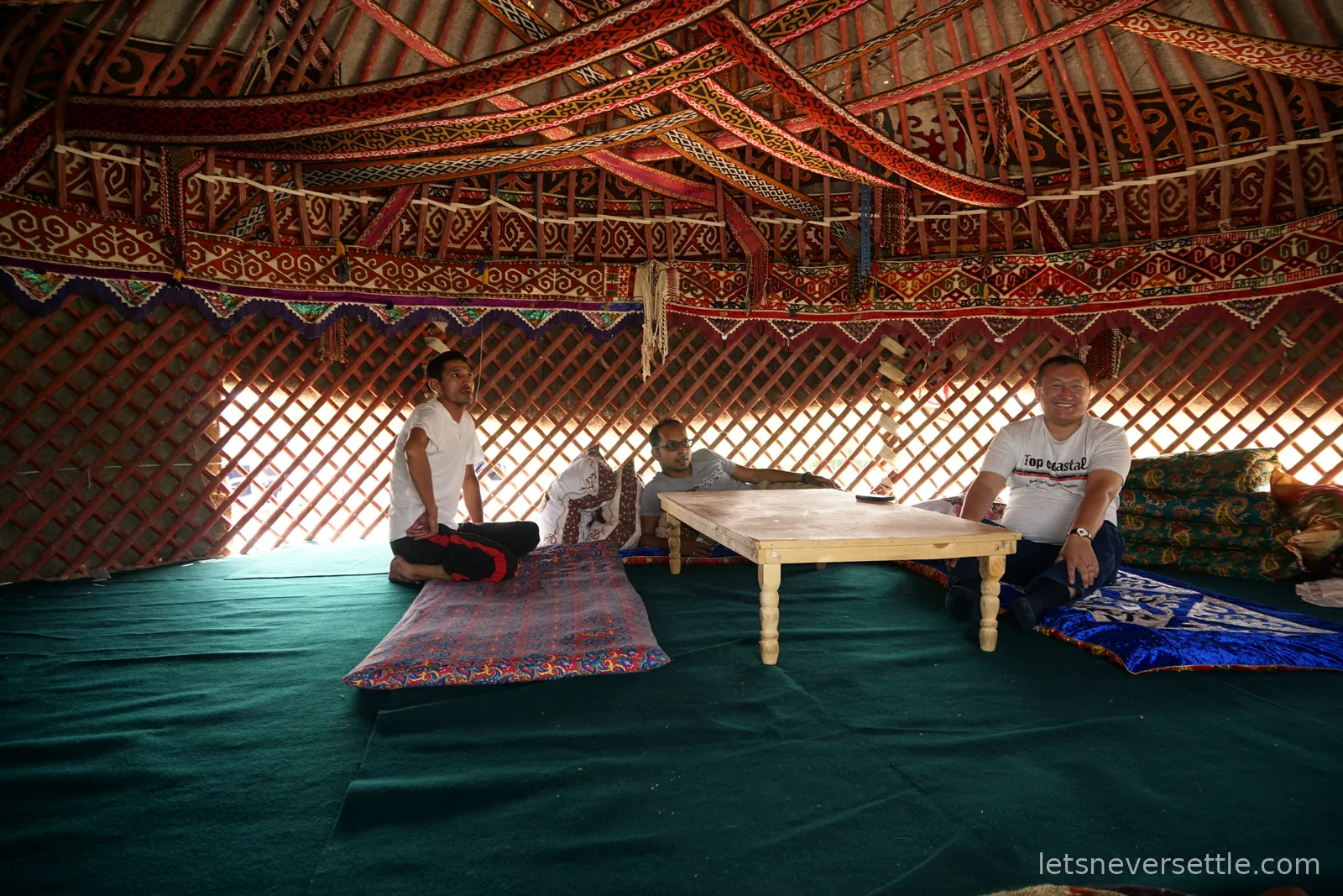 Inside a yurt, red lattice walls and roof poles hung with woven patterned bands, Dileep reclining at a low wooden table with two smiling hosts