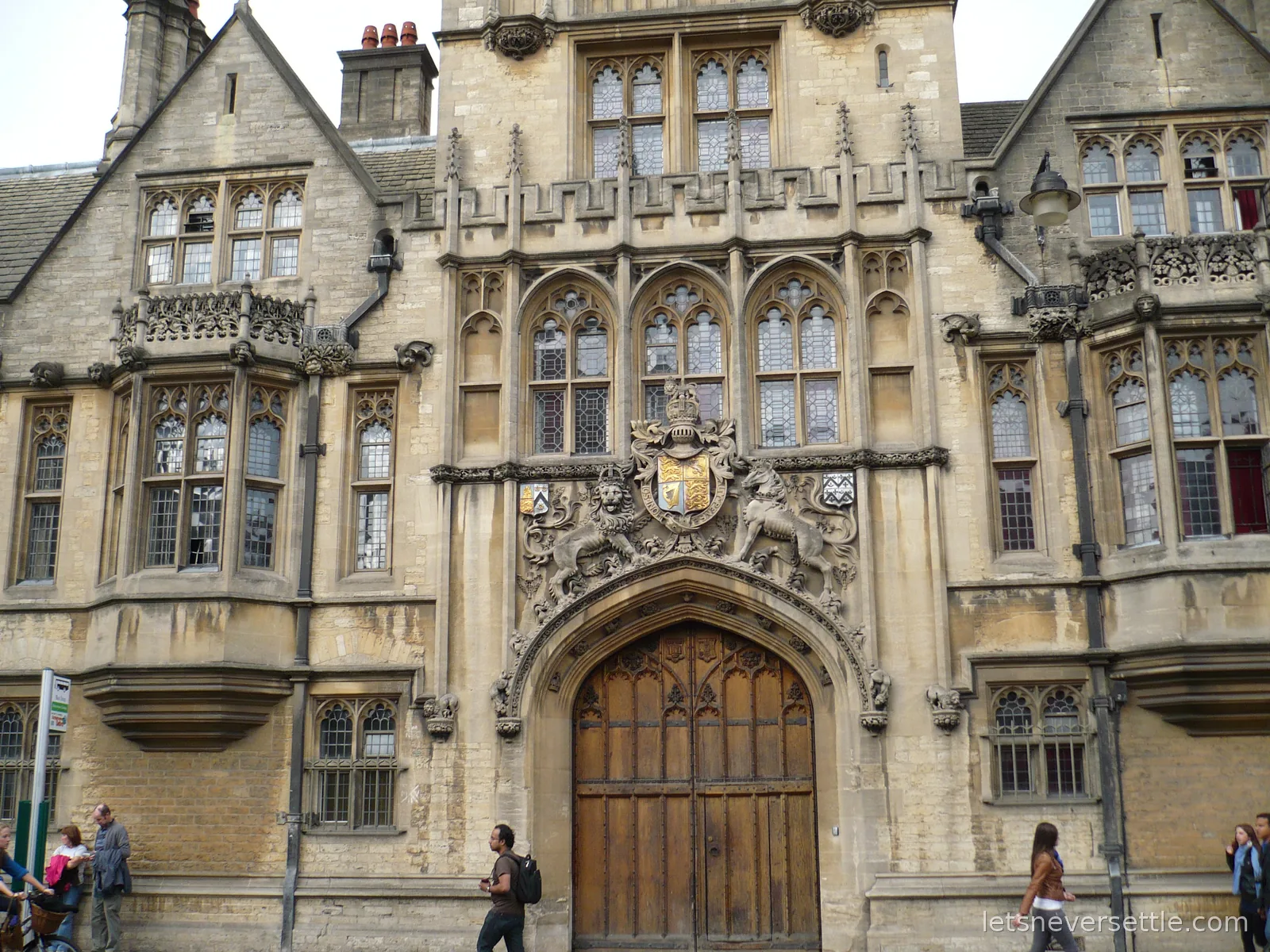A Gothic stone college gatehouse on Oxford's High Street with carved bay windows and a royal coat of arms above the wooden doors